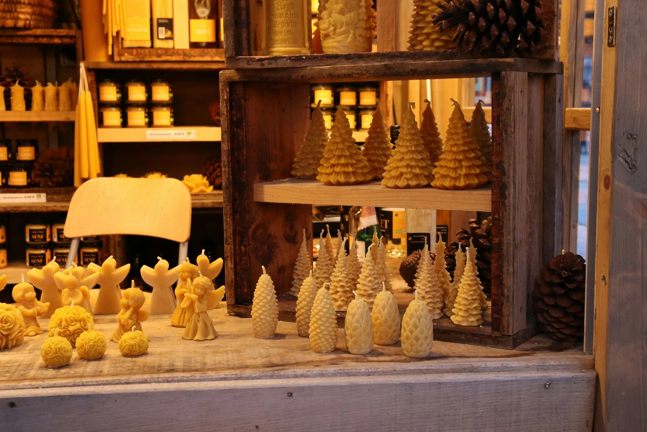 Display of multiple beeswax candles shaped like Christmas trees, angel figures, and decorative spheres at a market stall with shelves of yellow candles and pinecones in the background.