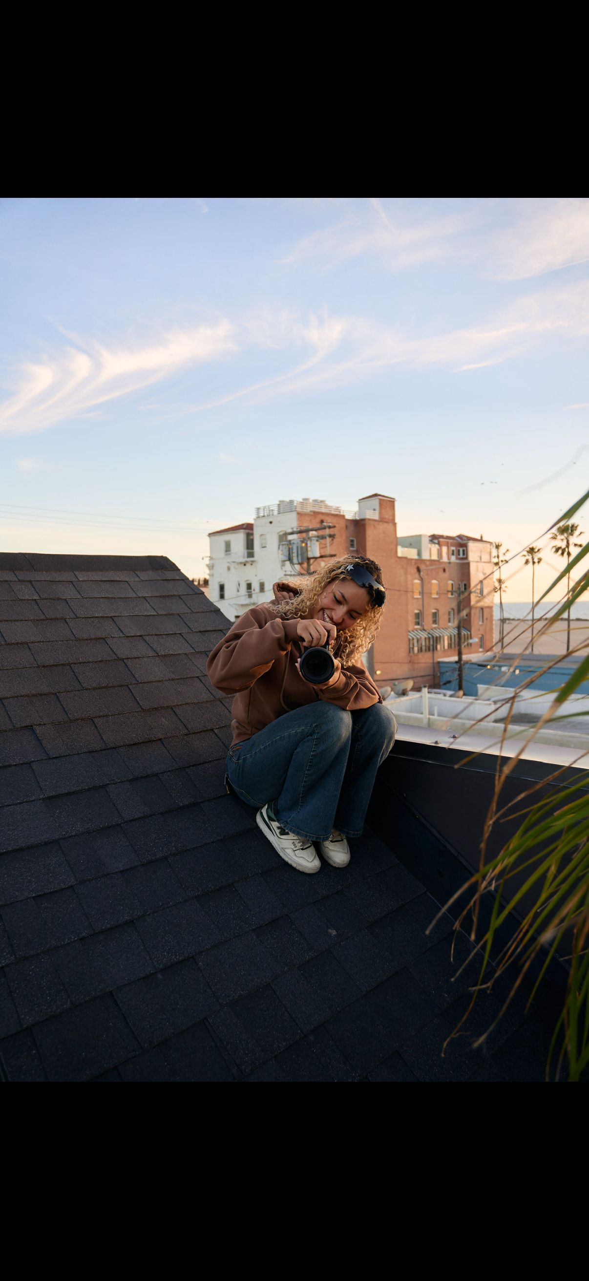 Young woman on rooftop using a camera during sunset with city buildings in background.
