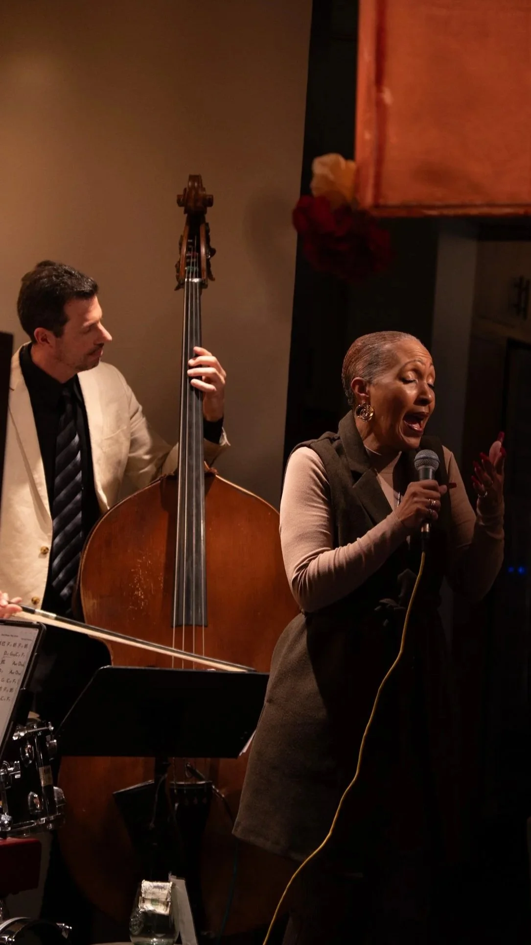 A woman singing into a microphone with a jazz band, including a man playing upright bass, performing in an intimate setting.