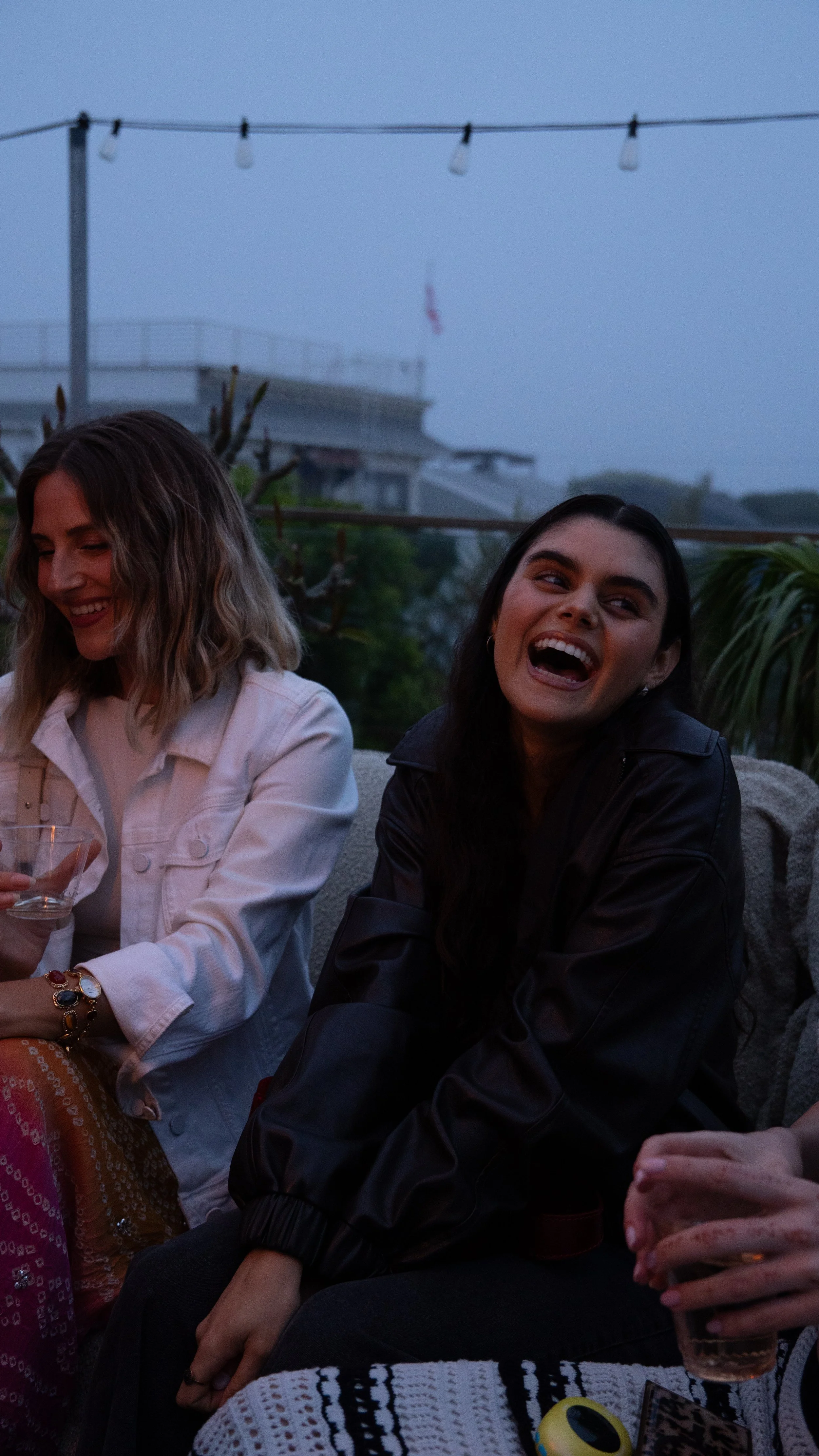 Two women sitting outdoors, laughing and enjoying drinks during evening or nighttime.