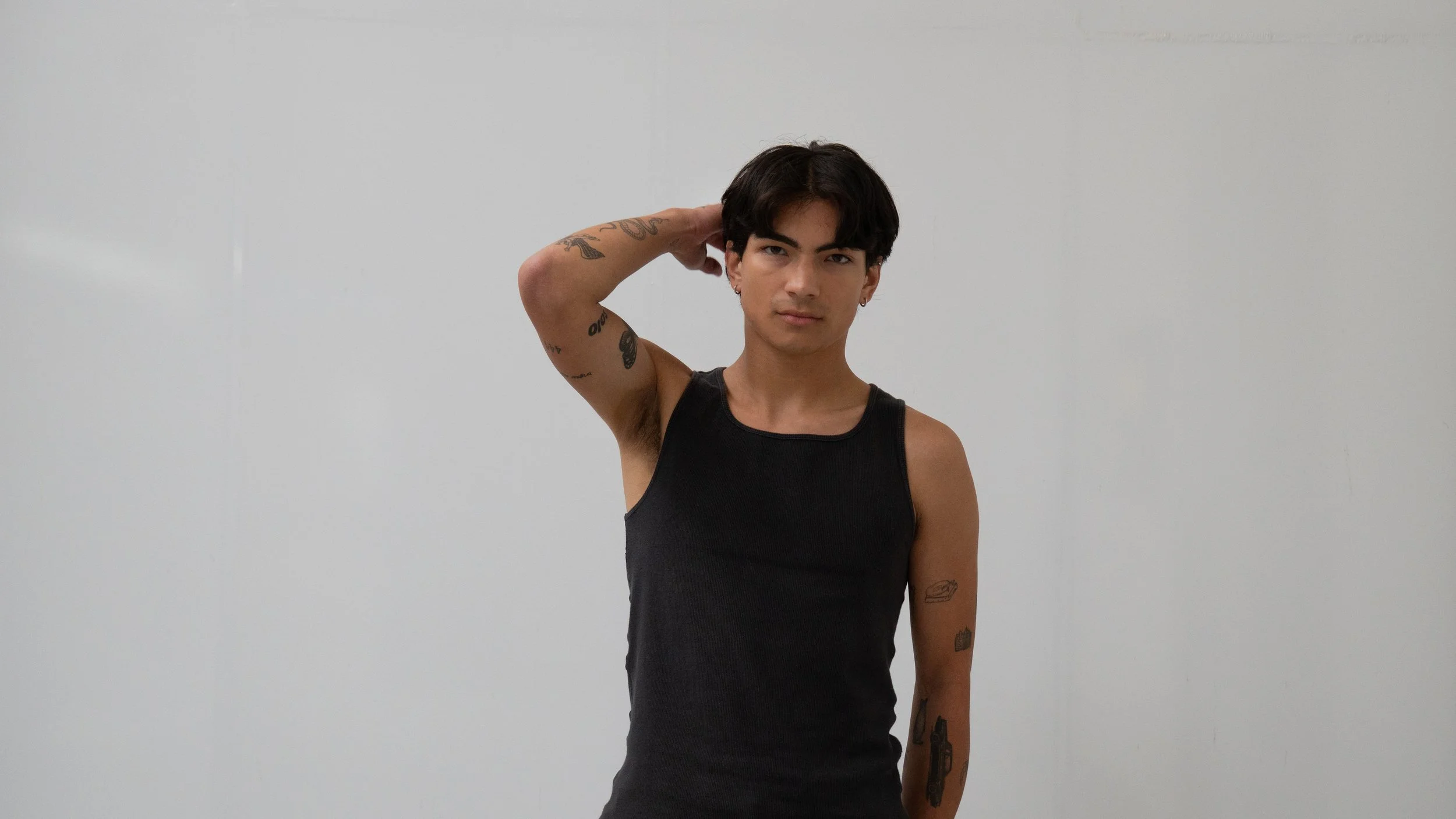 Outdoor Headshots Neutral. Young man with dark hair and tattoos wearing a black tank top, standing against a plain white wall, looking at the camera with one hand behind his head.