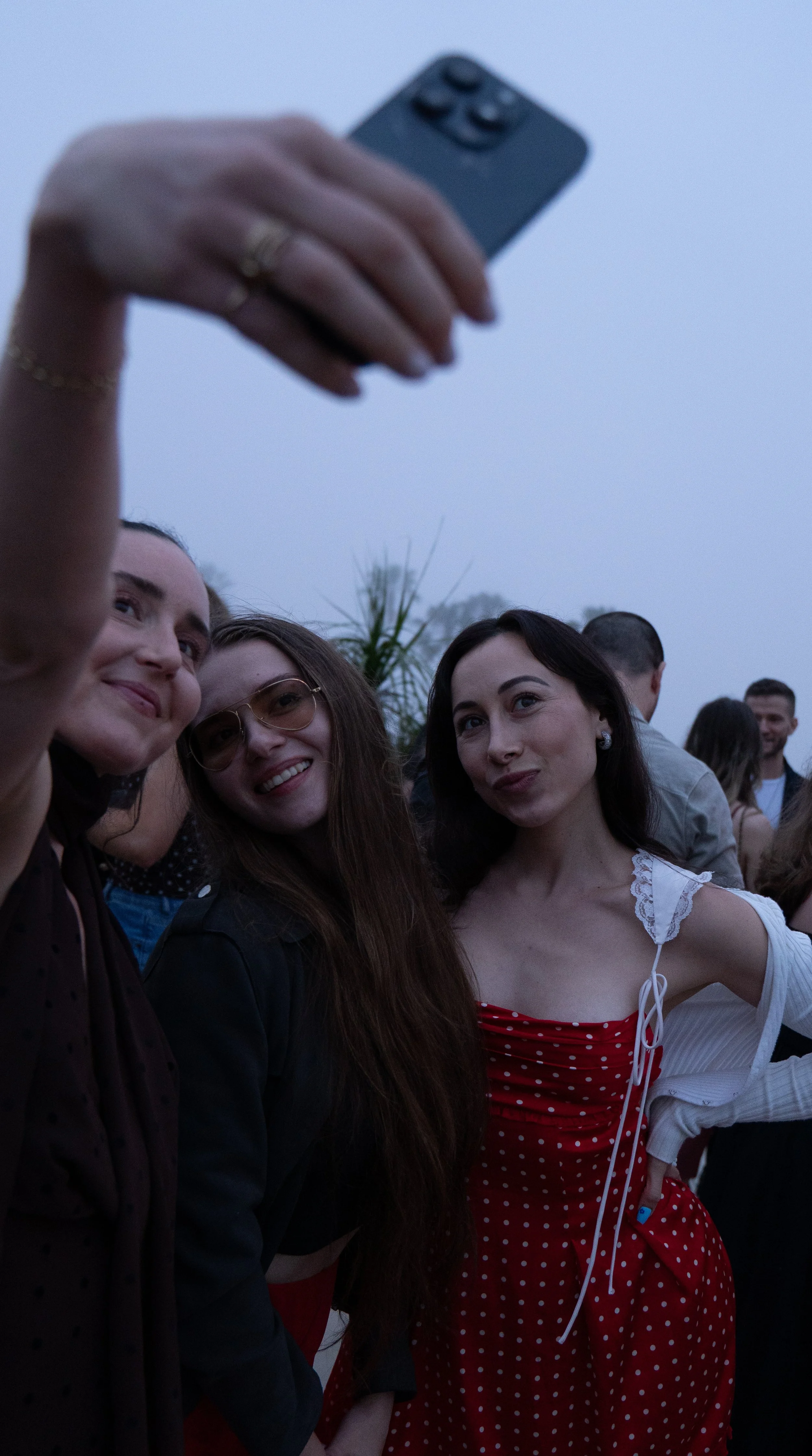 Event Photography. Three women taking a selfie at an outdoor event during dusk, with other people in the background.