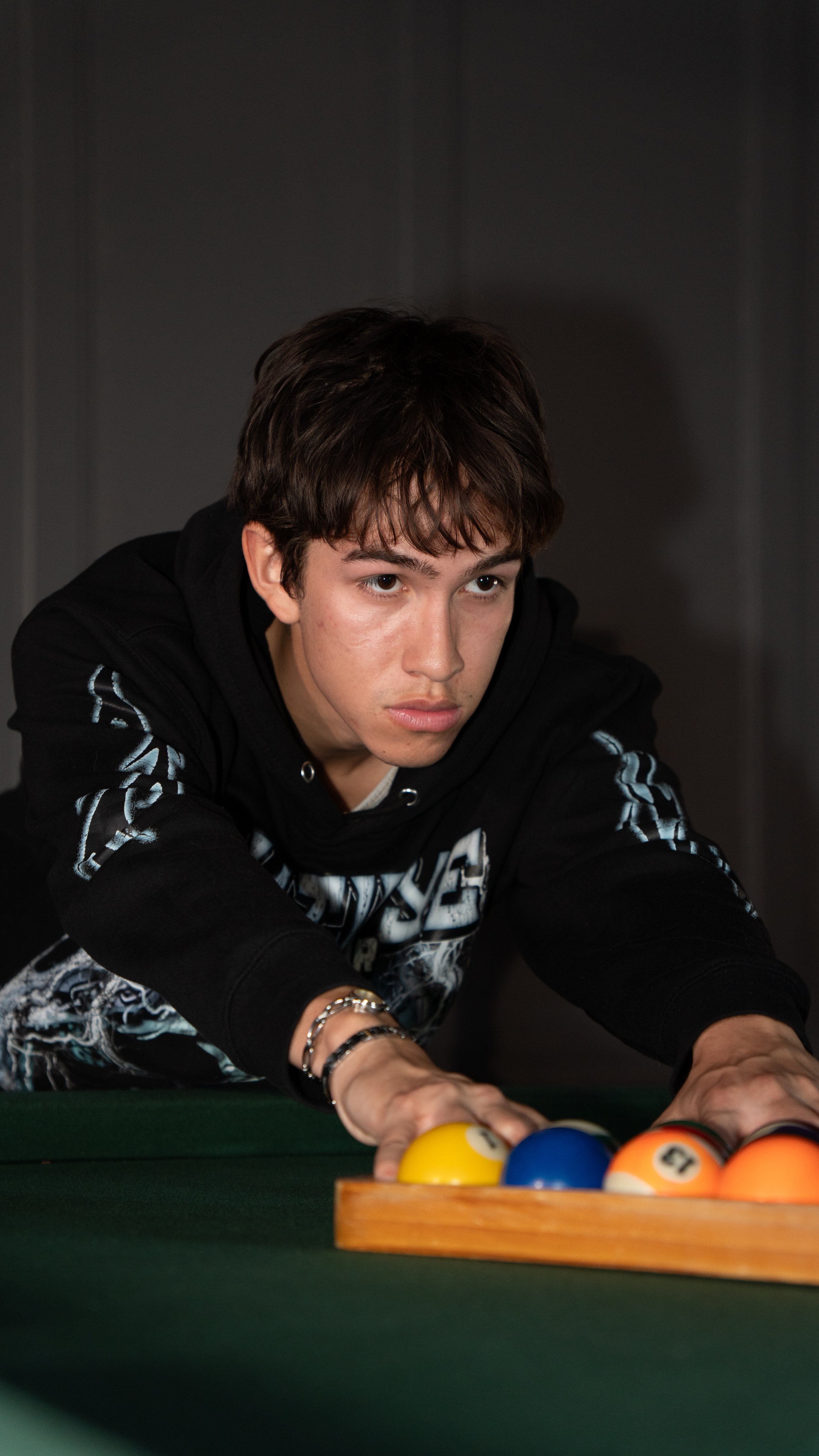 A young man with dark short hair wearing a black hoodie with white graphic design, leaning over a pool table.