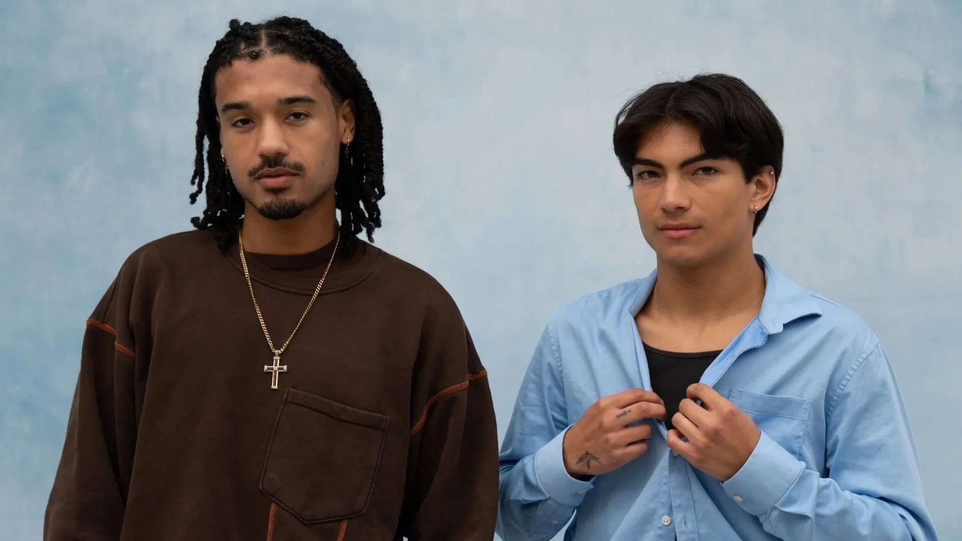 Two young men standing against a light blue background, one with long dreadlocks wearing a brown sweatshirt and a gold cross necklace, the other with short dark hair wearing a light blue button-up shirt and a black t-shirt underneath, adjusting the c