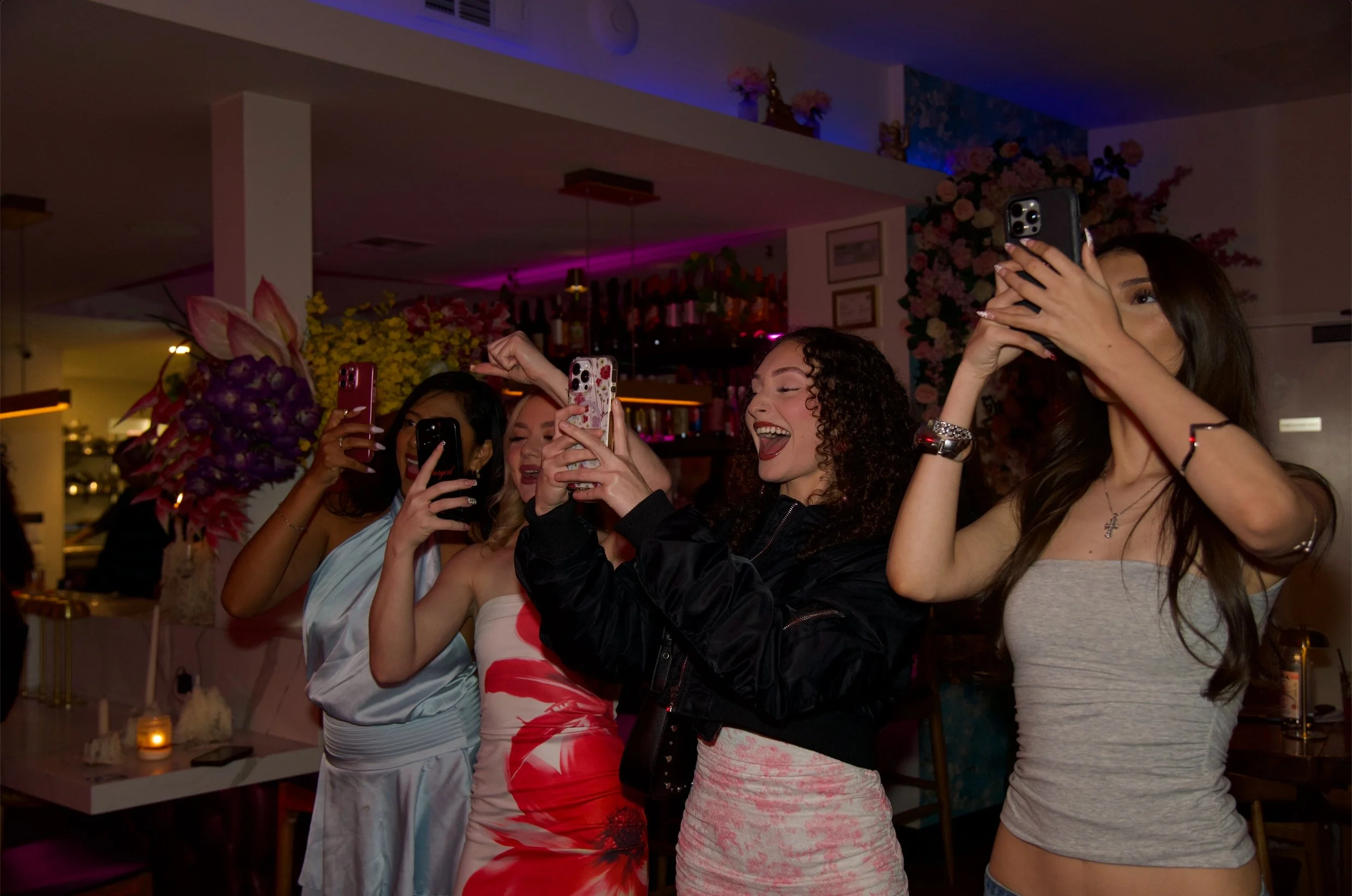 Four women at a party, taking selfies with their smartphones, in a decorated venue with flowers and ambient lighting.