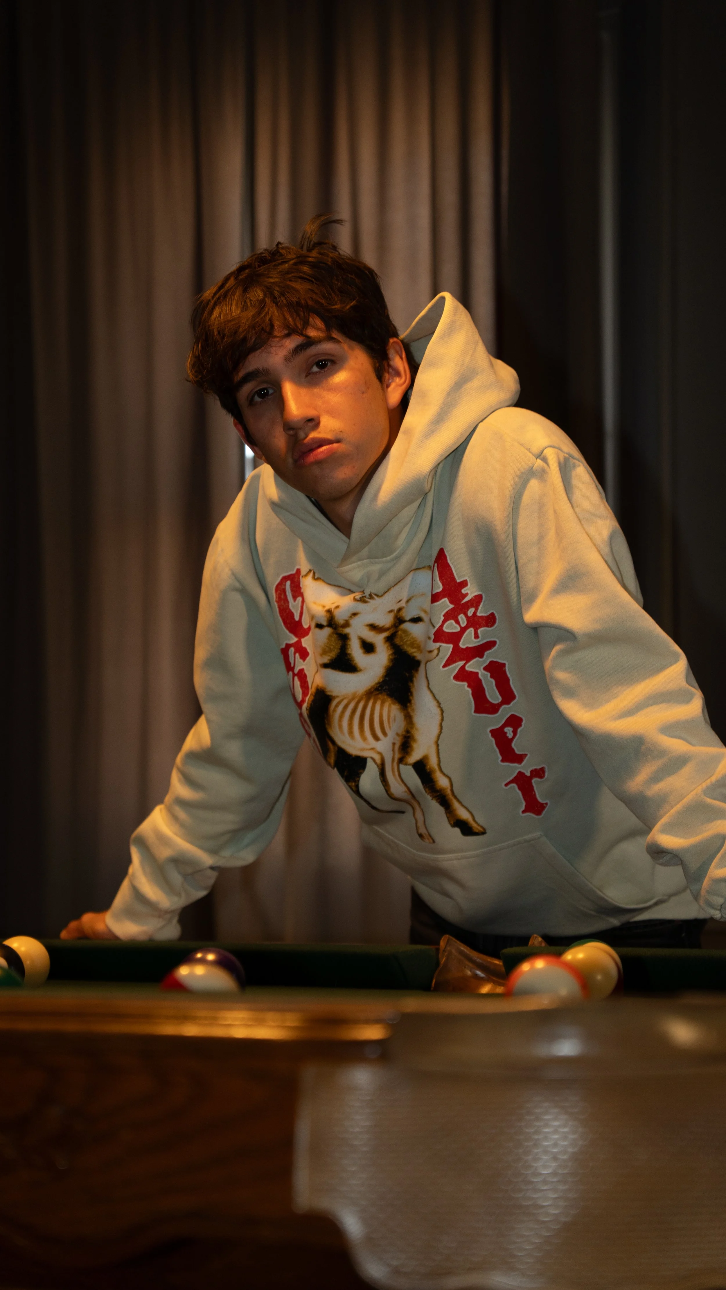 Product Photography, specifically modeled clothing images. Young man leaning over a pool table with a contemplative expression, wearing a cream-colored hoodie with a graphic of a tiger and red text.