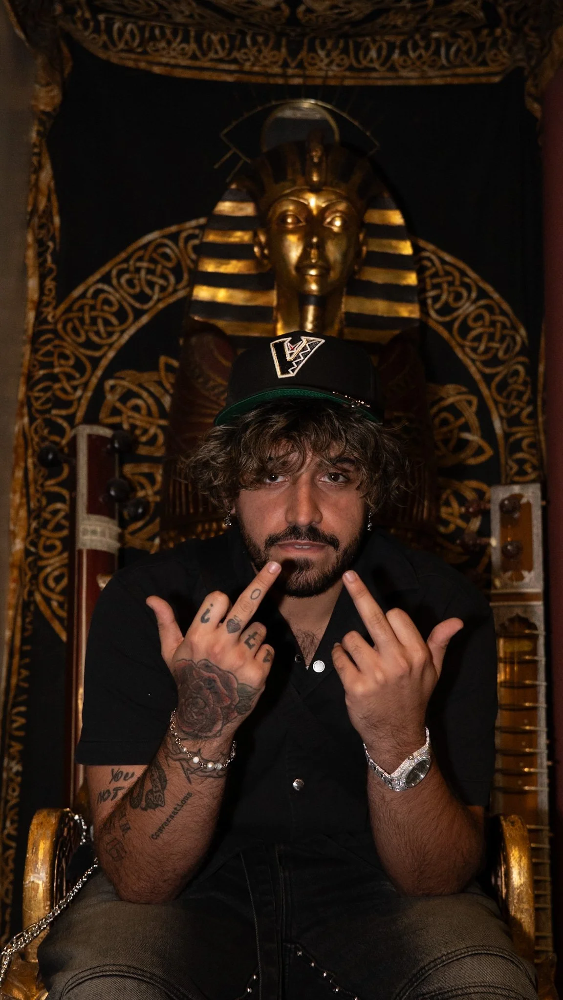 A man with tattoos and dark hair wearing a black baseball cap with a logo, sitting on a throne, making a rebellious gesture with his middle fingers raised. Behind him is a golden statue of a pharaoh's head and an ornate, dark decorative background.