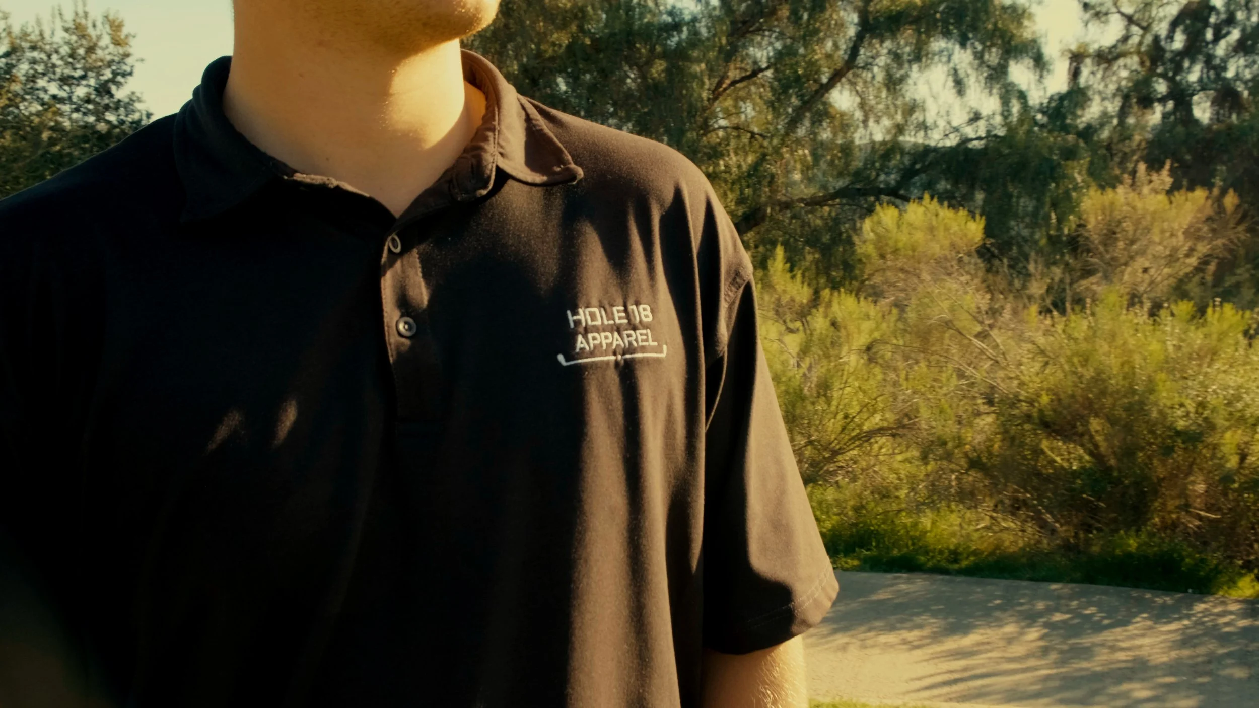 Close-up of a man wearing a black polo shirt with a logo that reads 'Holder8 Apparel', outdoors with trees and a road in the background.