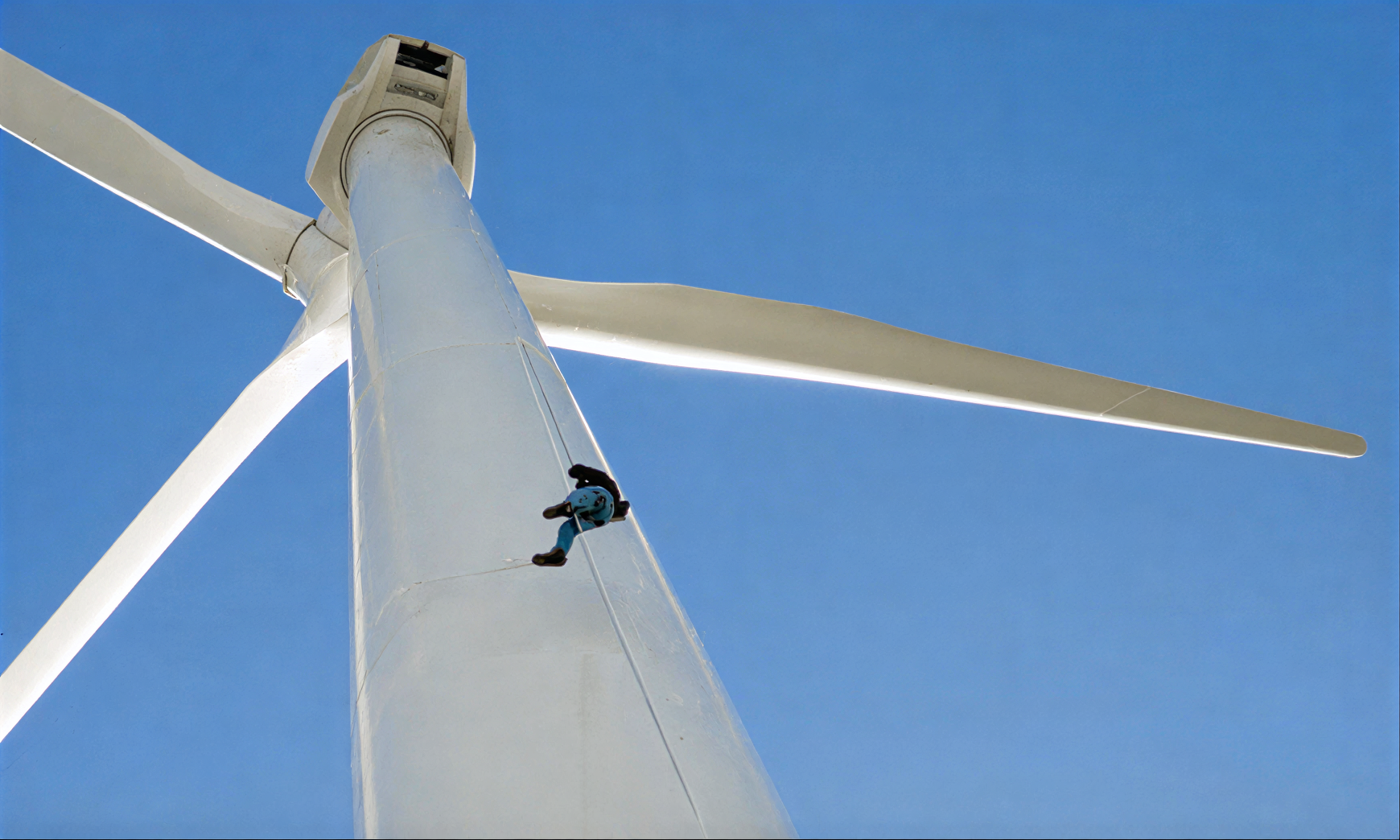 Close-up of a wind turbine tower with a maintenance worker rappelling down the side, set against a clear blue sky.