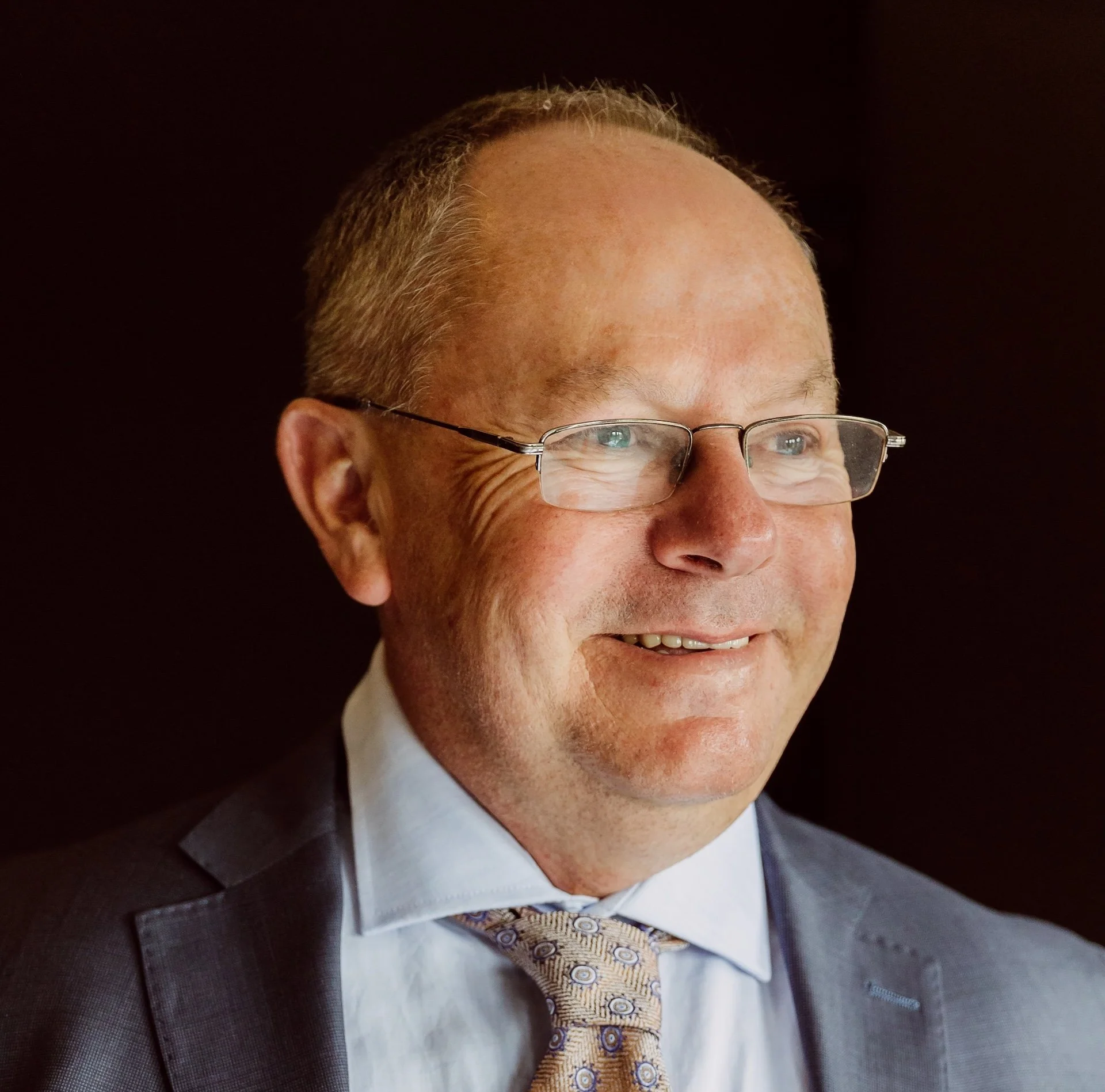 Close-up of a man with glasses, smiling, wearing a suit and tie, against a dark background.