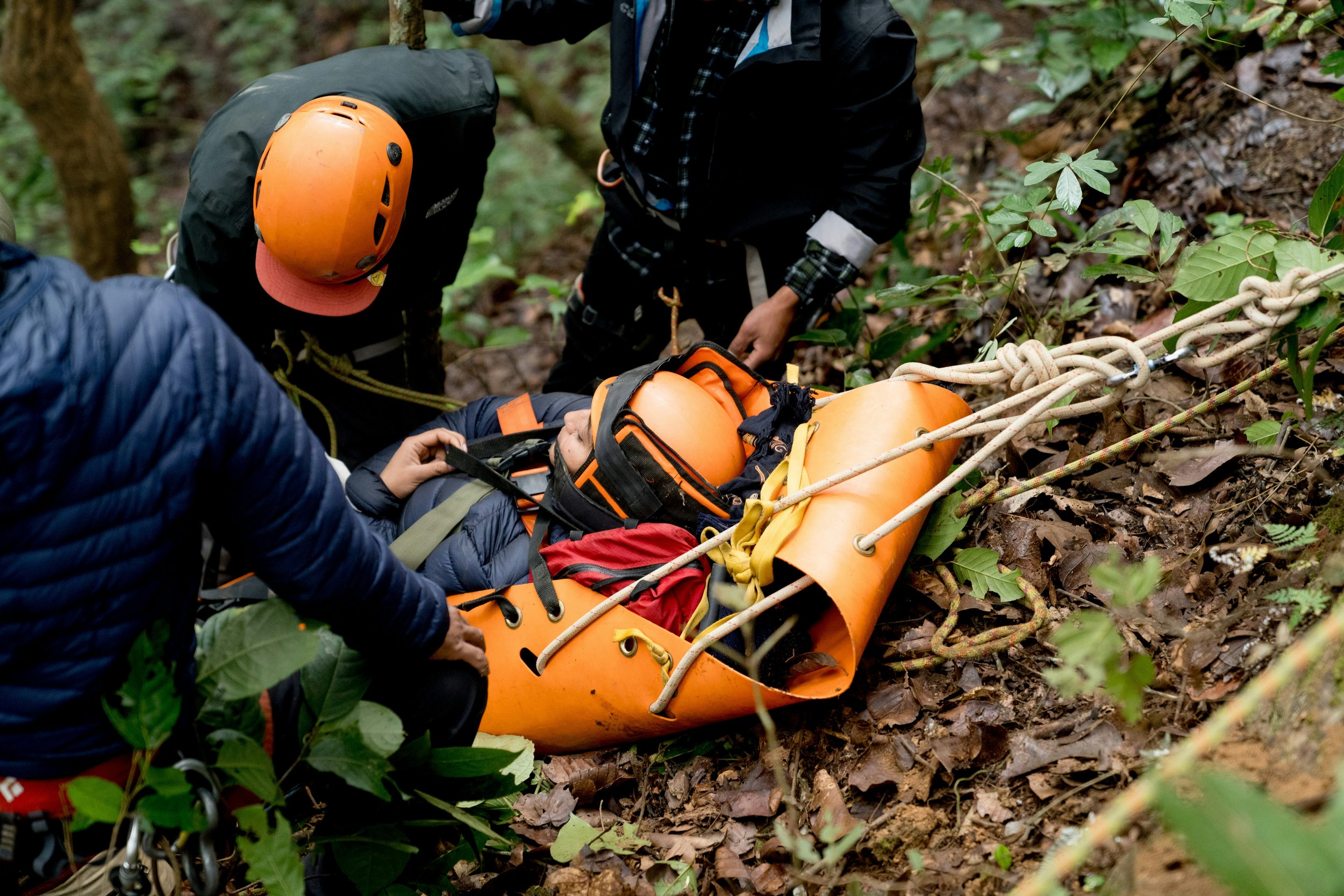 Rescue team uses a stretcher and ropes to assist a person lying on the forest ground, surrounded by trees and foliage.