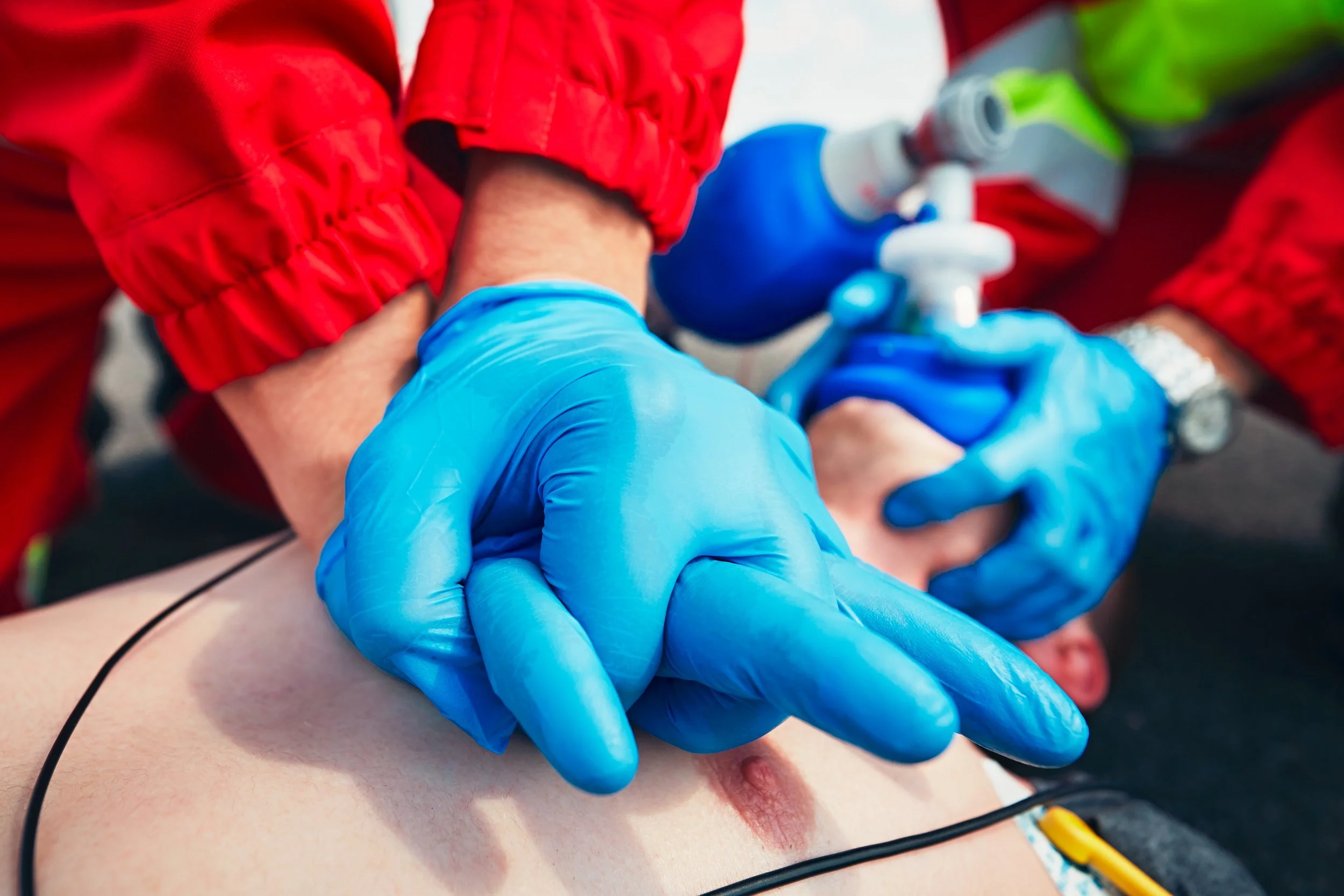 A healthcare professional wearing blue gloves and a red uniform performance CPR on a patient's chest, with medical equipment visible in the background.