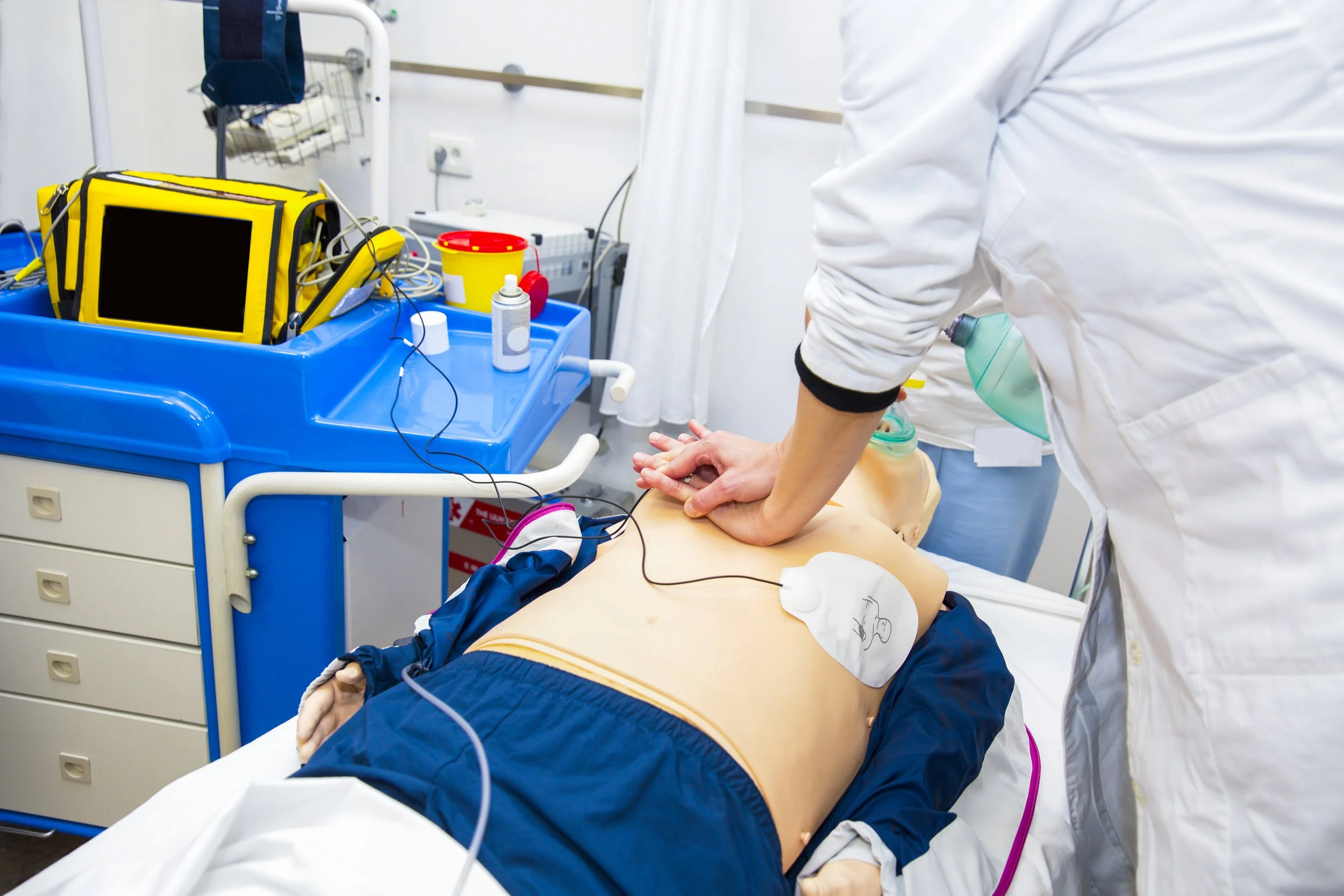 A healthcare professional performing CPR on a patient lying on a hospital bed in a medical room with various medical equipment and supplies.