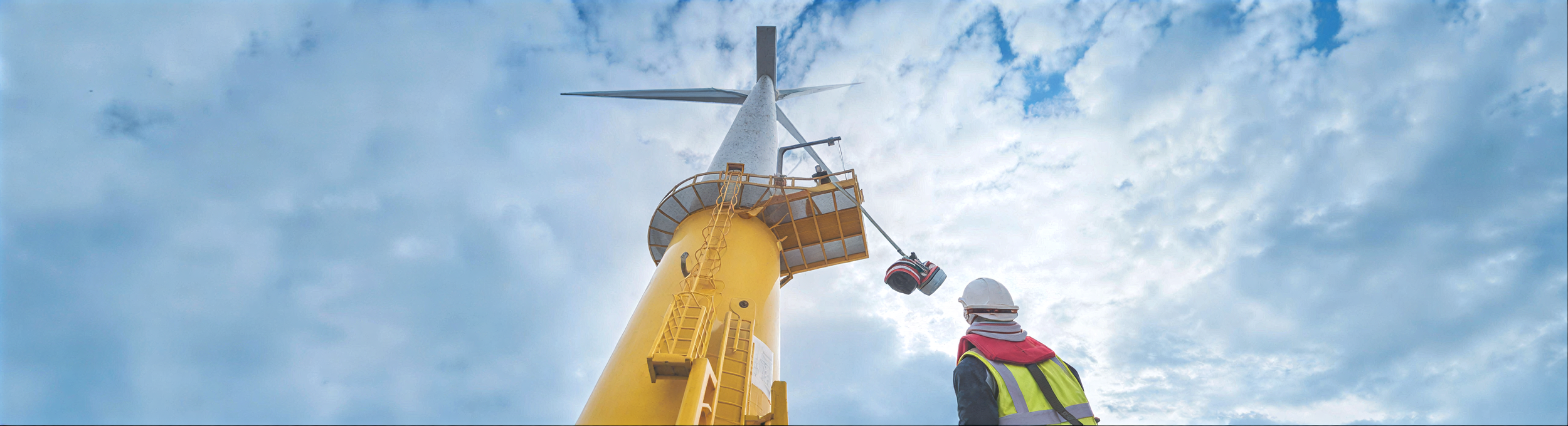 Worker in safety gear inspecting a large yellow wind turbine against a cloudy sky.