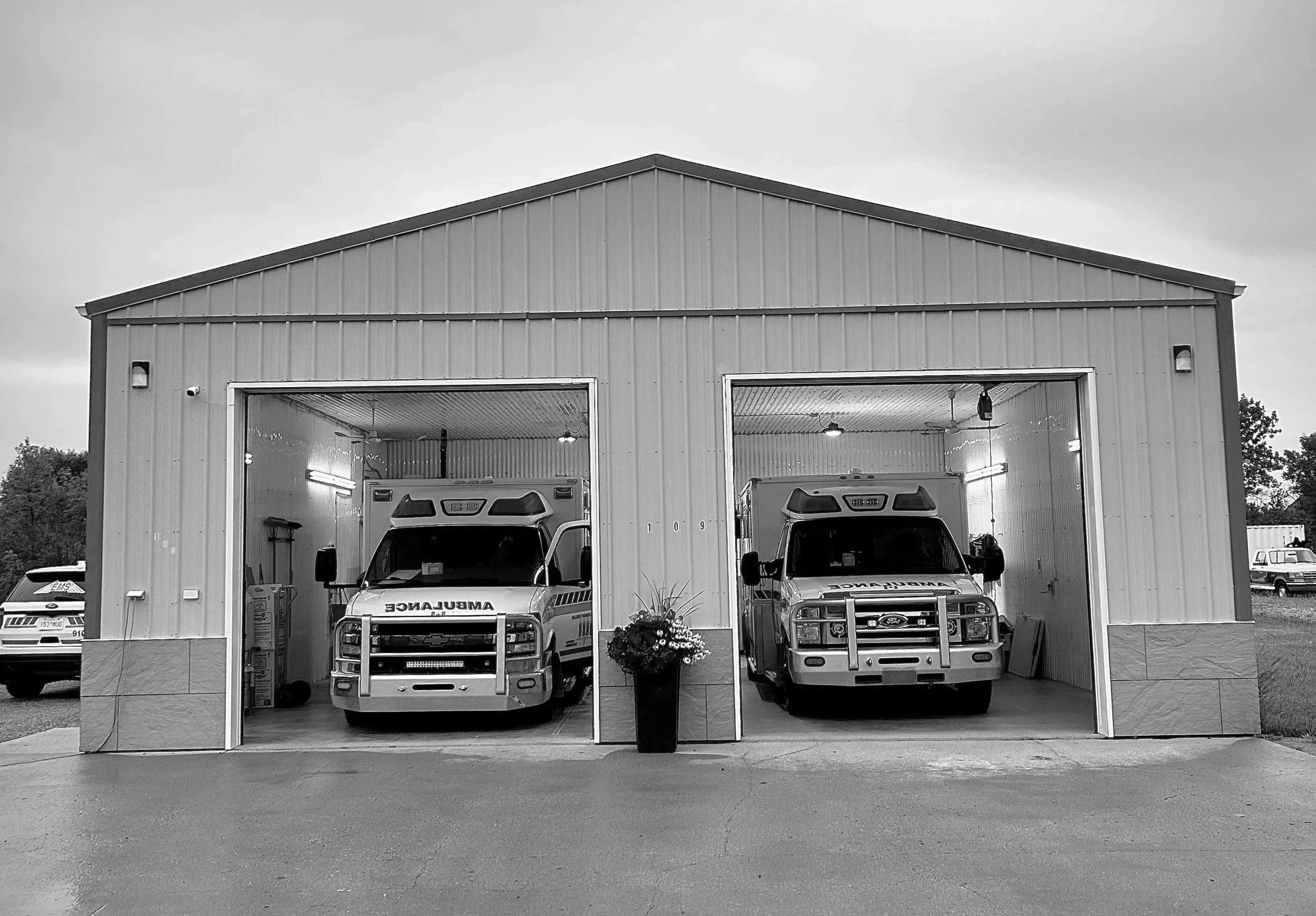 Black and white photo of a fire station with two fire trucks inside the garage bays. A potted plant is positioned between the two bays, and a police car is visible to the left outside.