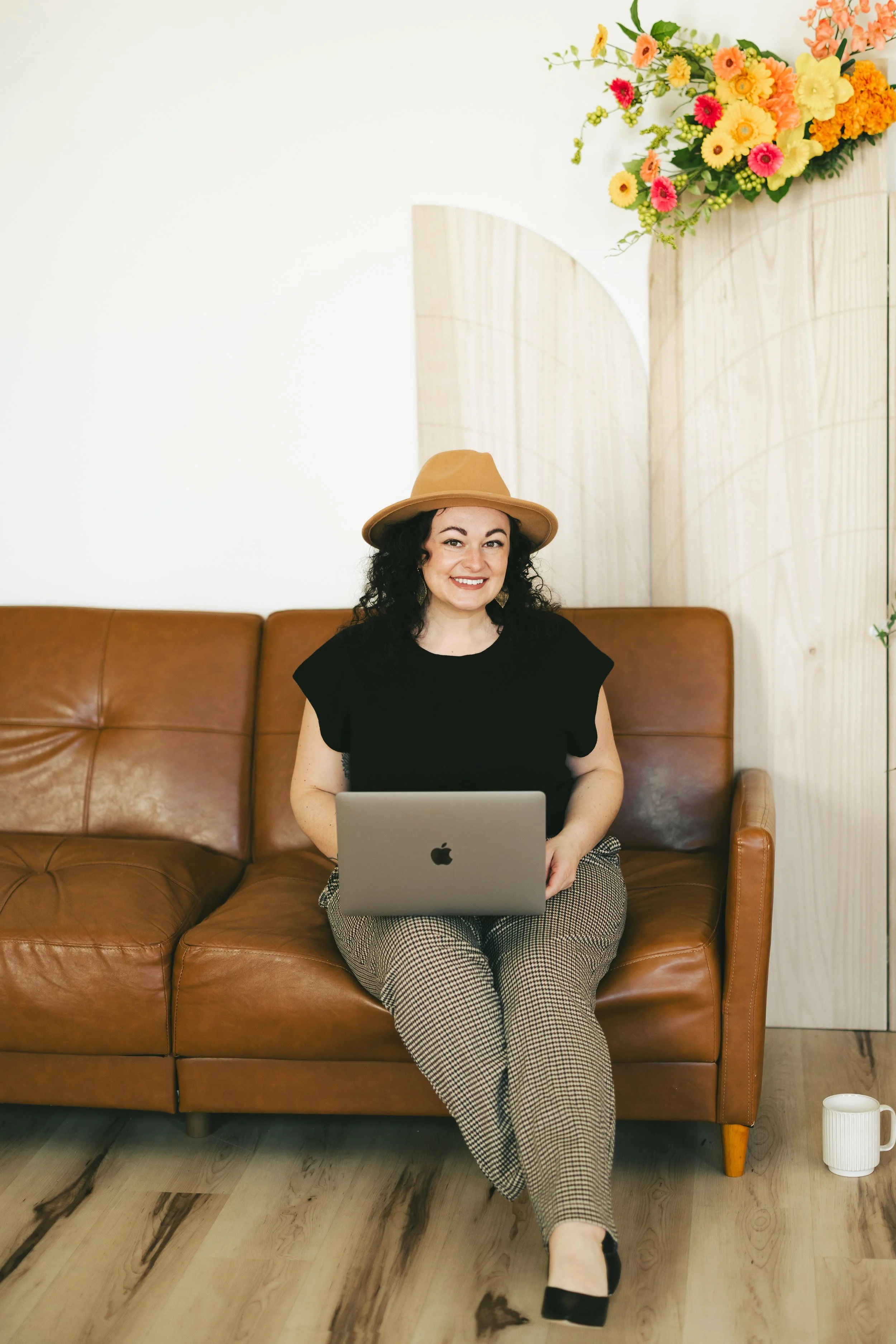 A woman sitting on a brown leather couch with a laptop on her lap, smiling at the camera, wearing a tan hat, black top, and checkered pants, in a room with light-colored wooden flooring and a floral arrangement on a wooden partition in the background.