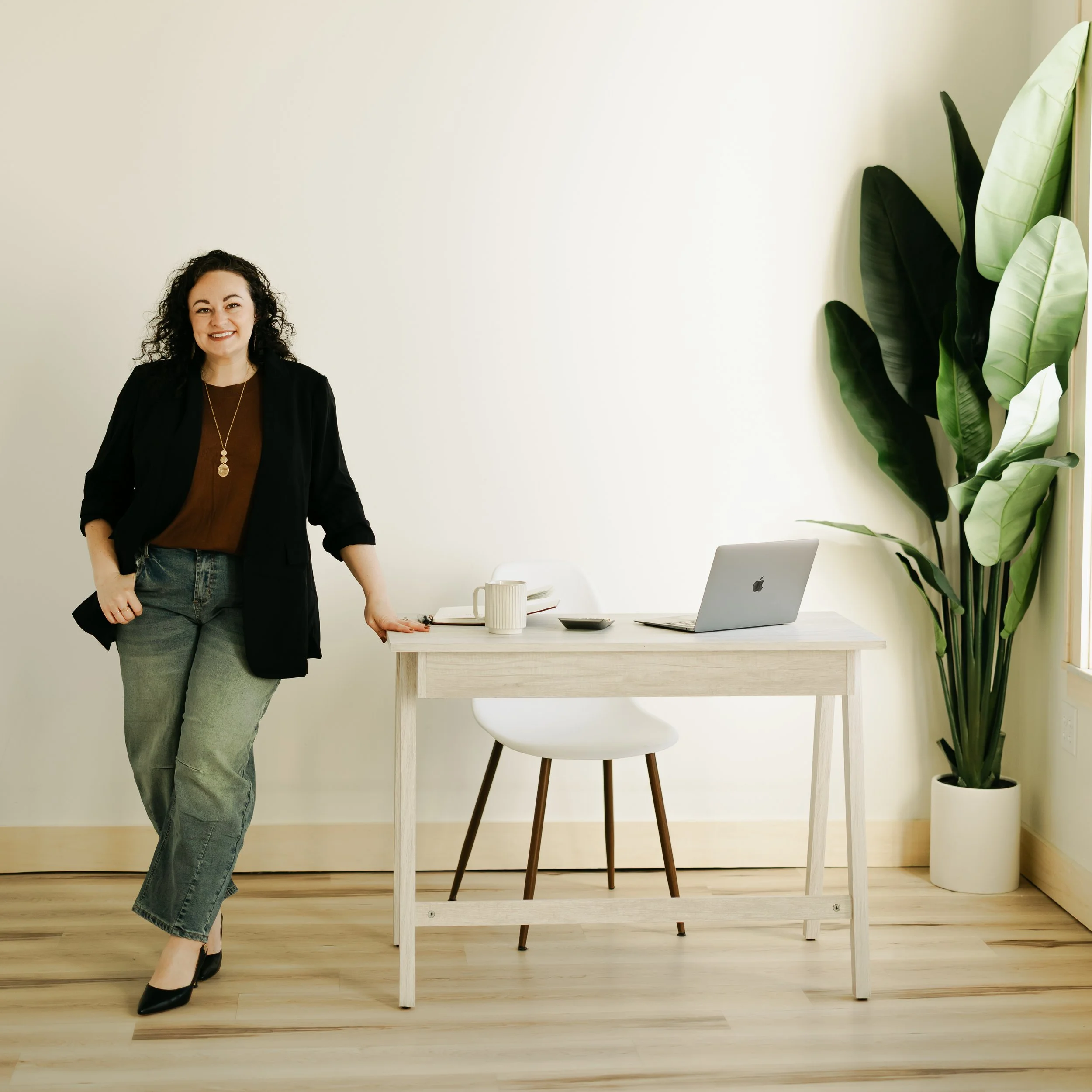 A woman with curly dark hair, wearing a black blazer, brown top, light blue jeans, and black high heels, standing next to a minimalist white desk with a silver laptop, a white coffee mug, and a notebook with a pen, against a plain off-white wall with large green tropical plant leaves on the right.