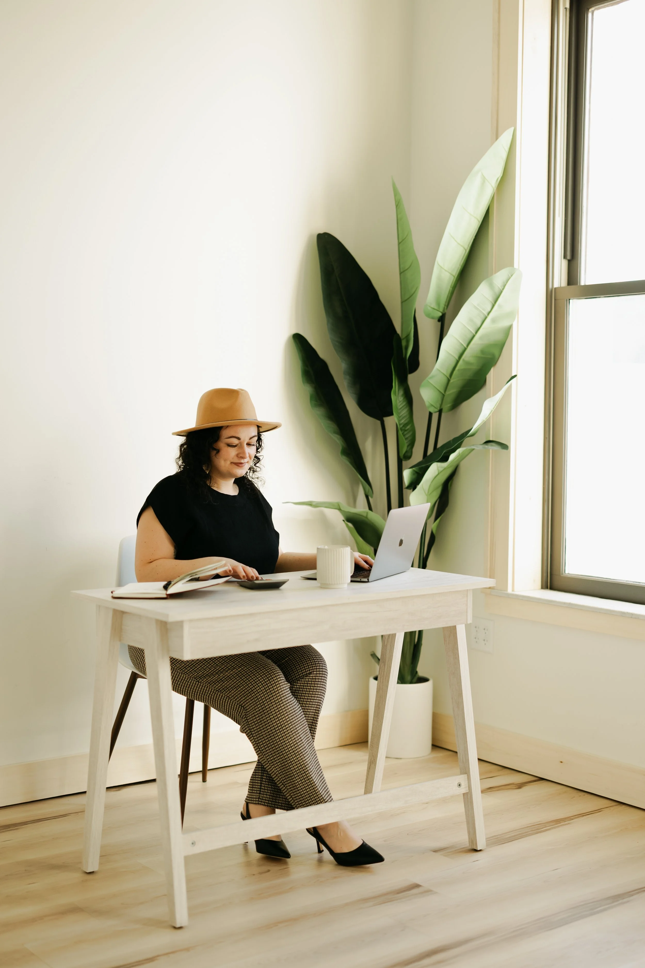 Woman sitting at a light wooden desk, working on a laptop, with a large green plant next to her near a window.
