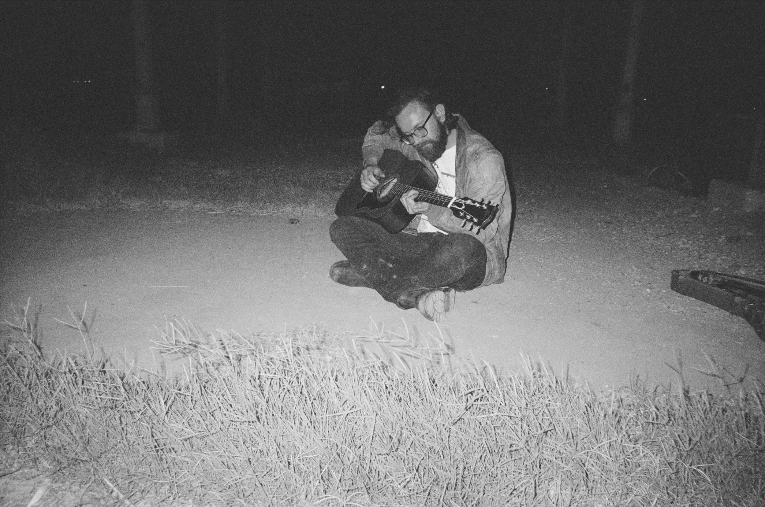A man with glasses and a beard, sitting cross-legged on the ground at night, playing an acoustic guitar, with grass and a backpack nearby.
