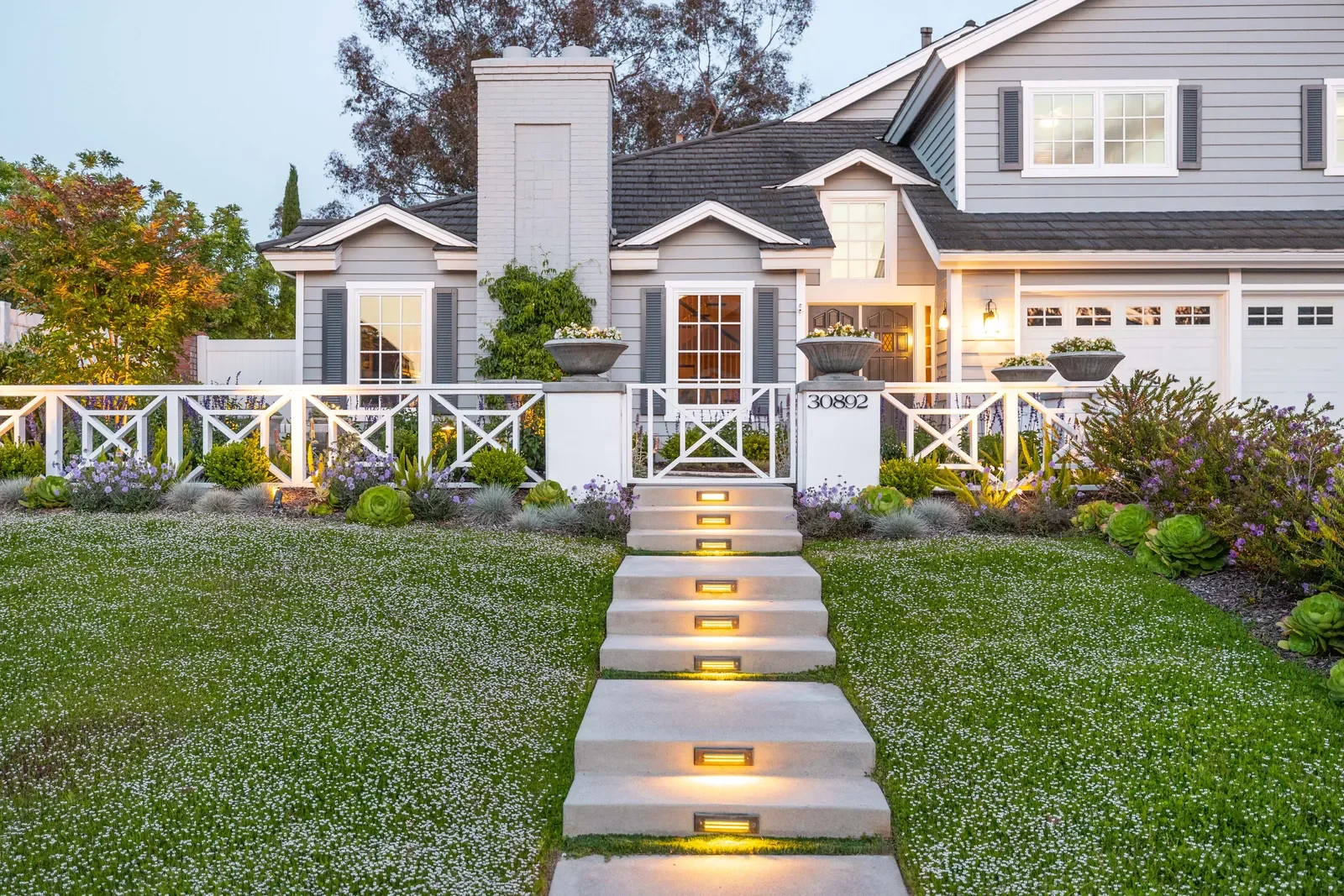 Front yard of a modern house with a landscaped garden, illuminated pathway steps, shrubs, and a white fence, taken at dusk.