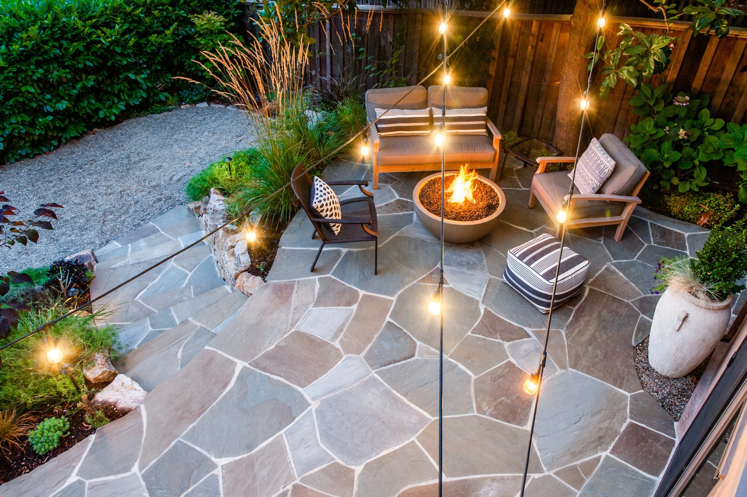 An outdoor backyard patio with stone flooring, surrounded by greenery and a wooden fence, featuring patio furniture arranged around a fire pit, with string lights hanging above.