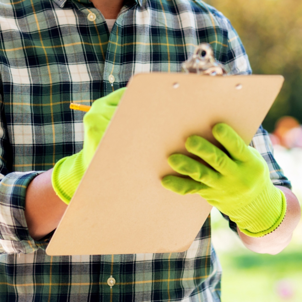 Close-up of a person wearing a plaid shirt and bright green gloves, holding a clipboard and writing with a pencil outdoors.
