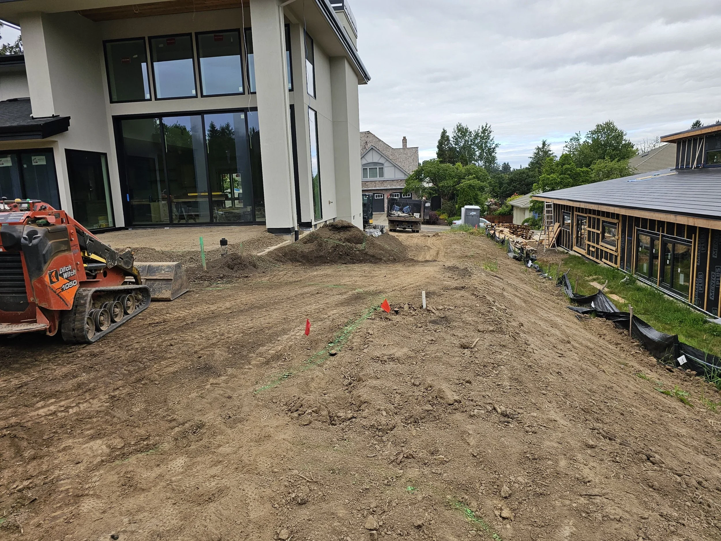 A residential backyard under construction with dirt and earthwork. There is a modern house with large glass windows on the left, and a partially constructed building with scaffolding and black sheathing on the right. Construction equipment, including a mini excavator, and various construction materials are visible.
