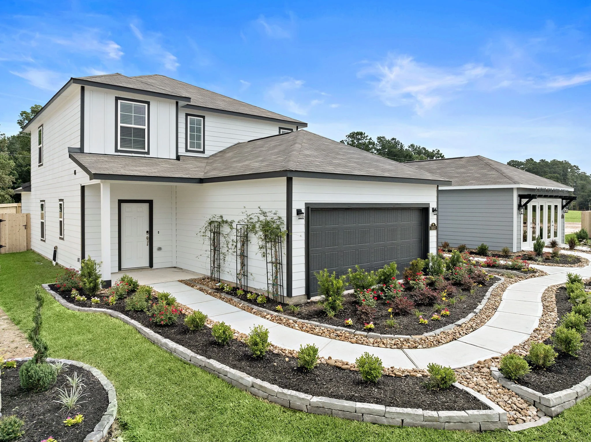 Front yard of a modern two-story house with white and gray siding, surrounded by landscaped flower beds, small shrubs, and a paved walkway under a blue sky.