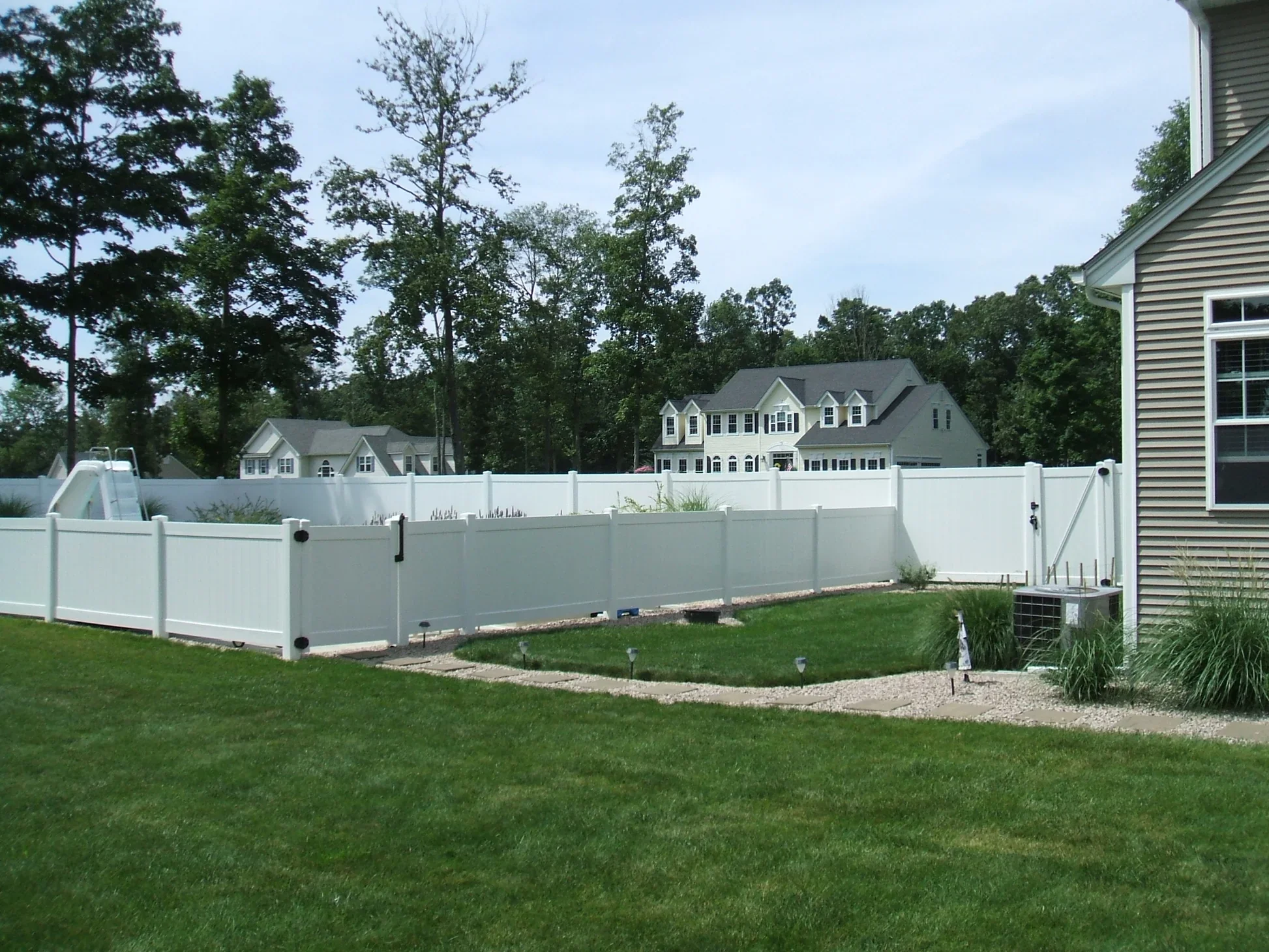 Residential backyard with a white privacy fence, green lawn, and a gravel pathway. A house with beige siding and windows is visible on the right. Several tall trees and neighboring houses are in the background under a blue sky.