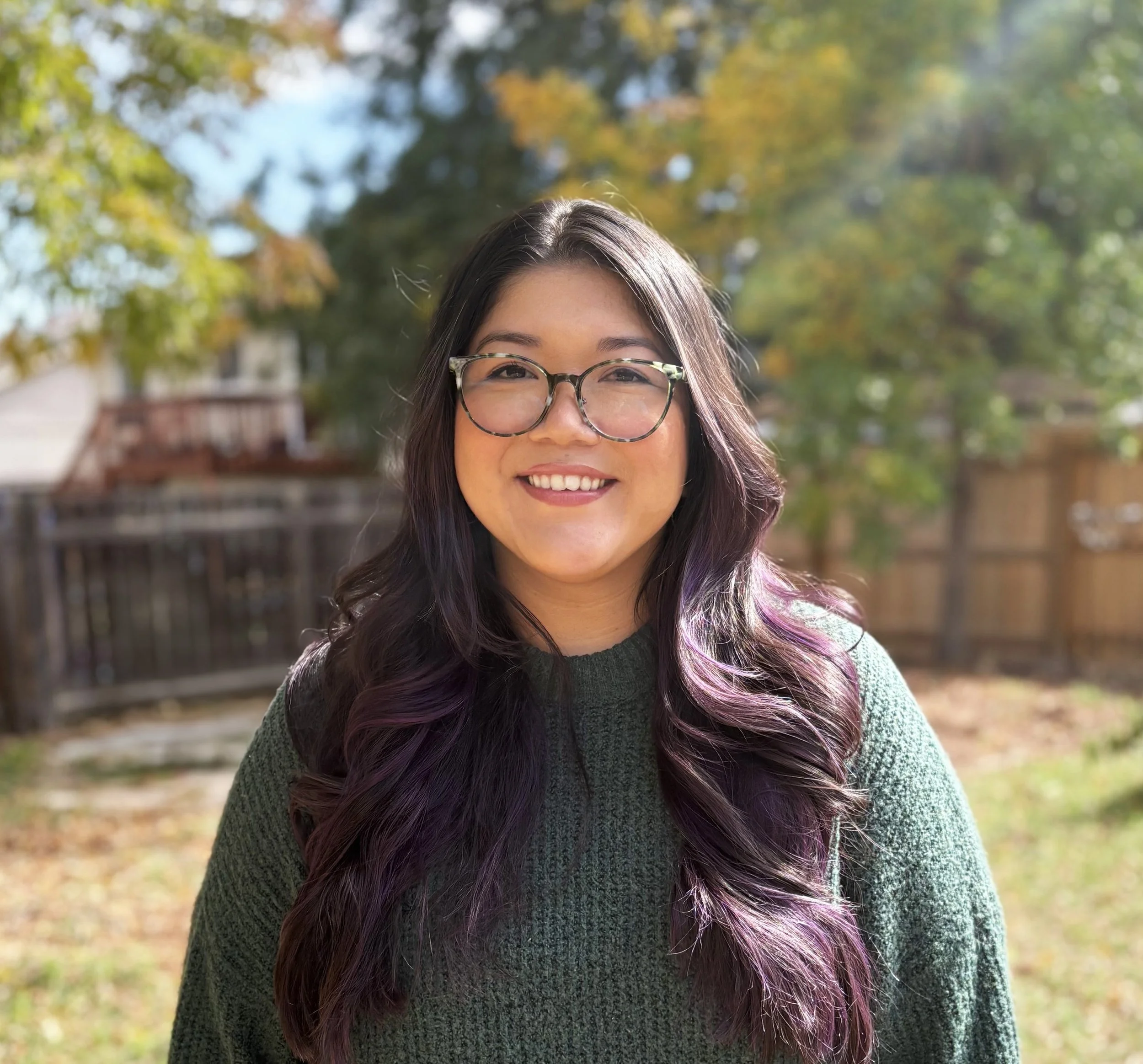 A woman with long, wavy hair that has purple tips, wearing glasses and a knitted green sweater, smiling outdoors with trees in the background.