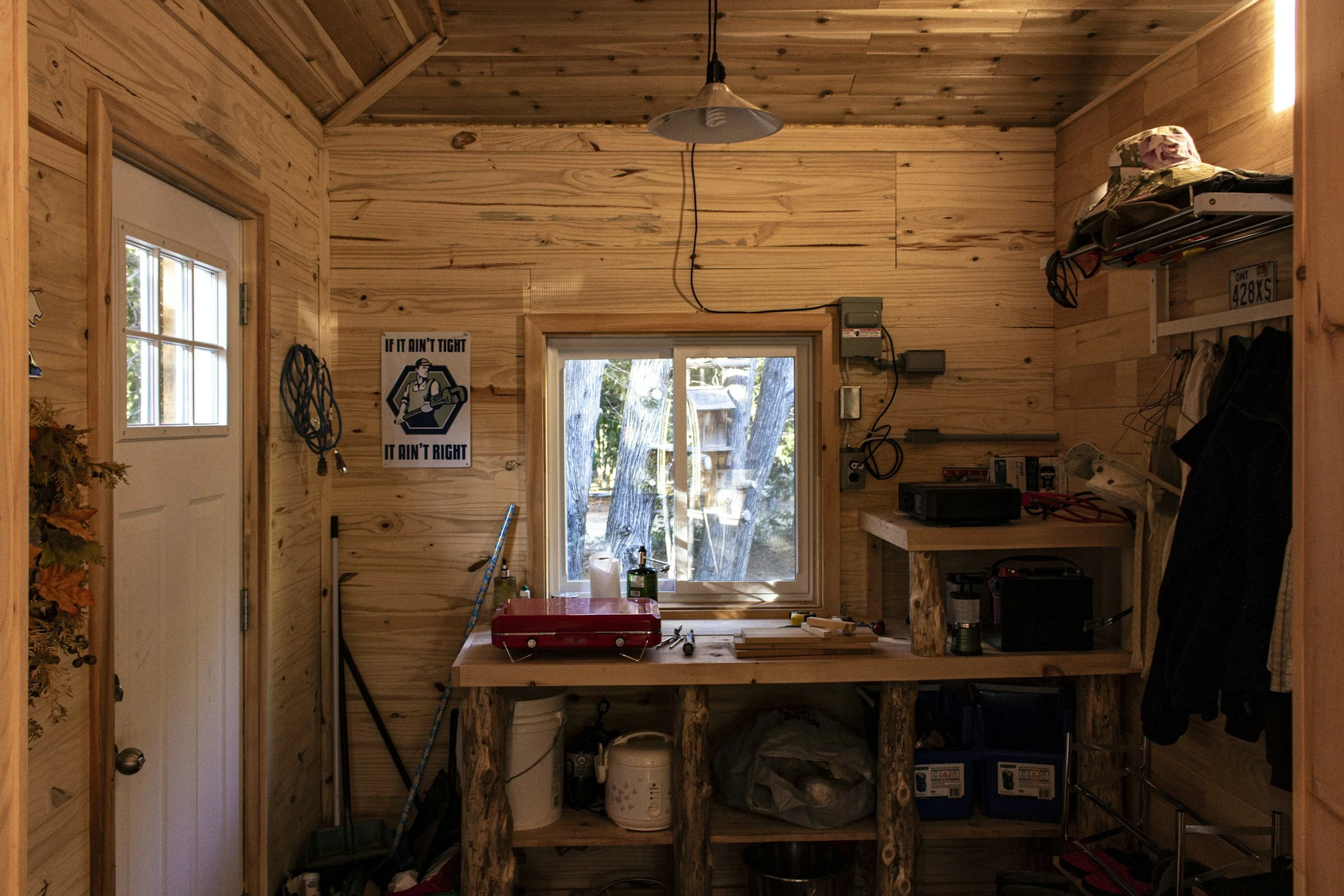 A small wood-paneled workshop with a window looking outside, a workbench with tools, a lamp hanging from the ceiling, and various supplies storage. A poster on the wall reads 'If it ain't tight, it ain't right'.
