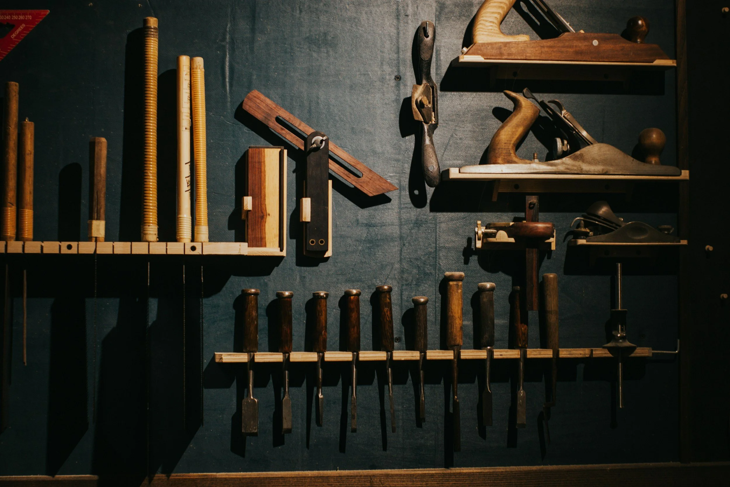 A wall mounted display of woodworking tools, including chisels, hand saws, and planes, organized on wooden racks.