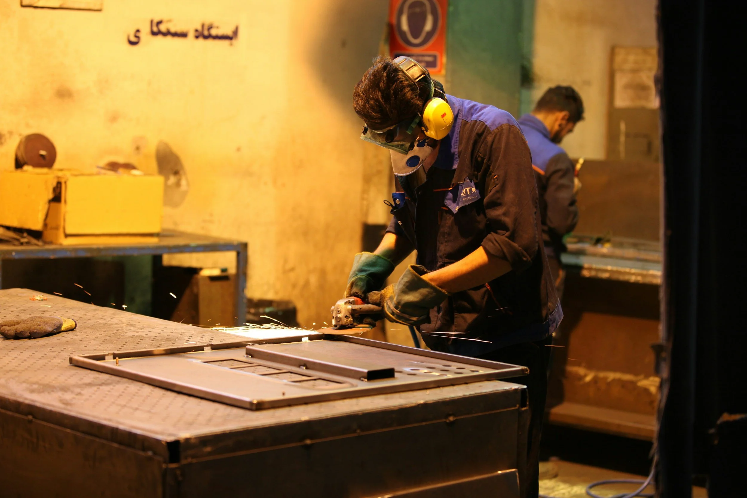A worker in safety gear using an angle grinder to cut or grind metal, with sparks flying, in a workshop or industrial setting.