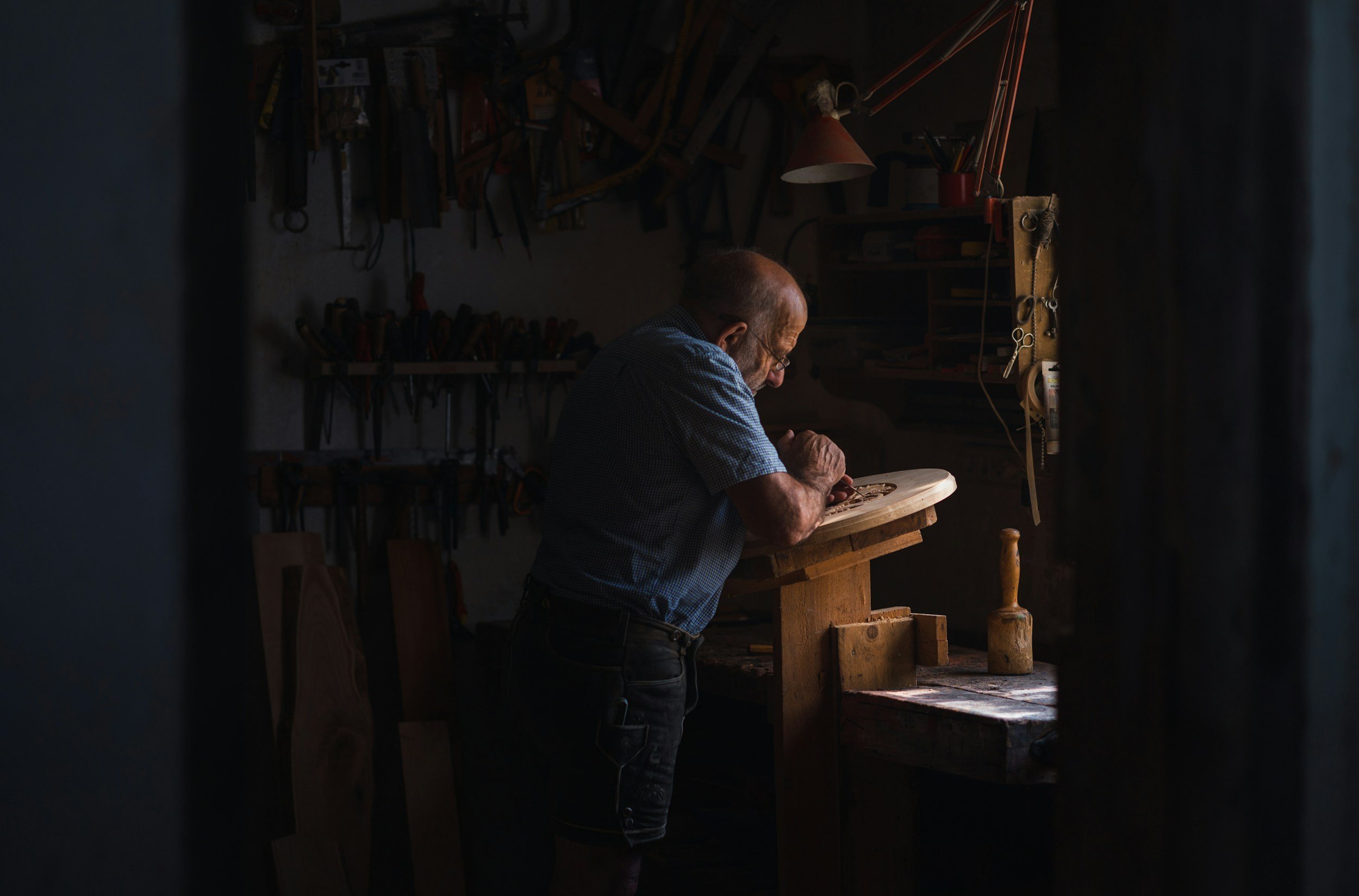 An elderly man working in a dimly lit woodworking workshop, carving on a piece of wood placed on a workbench.