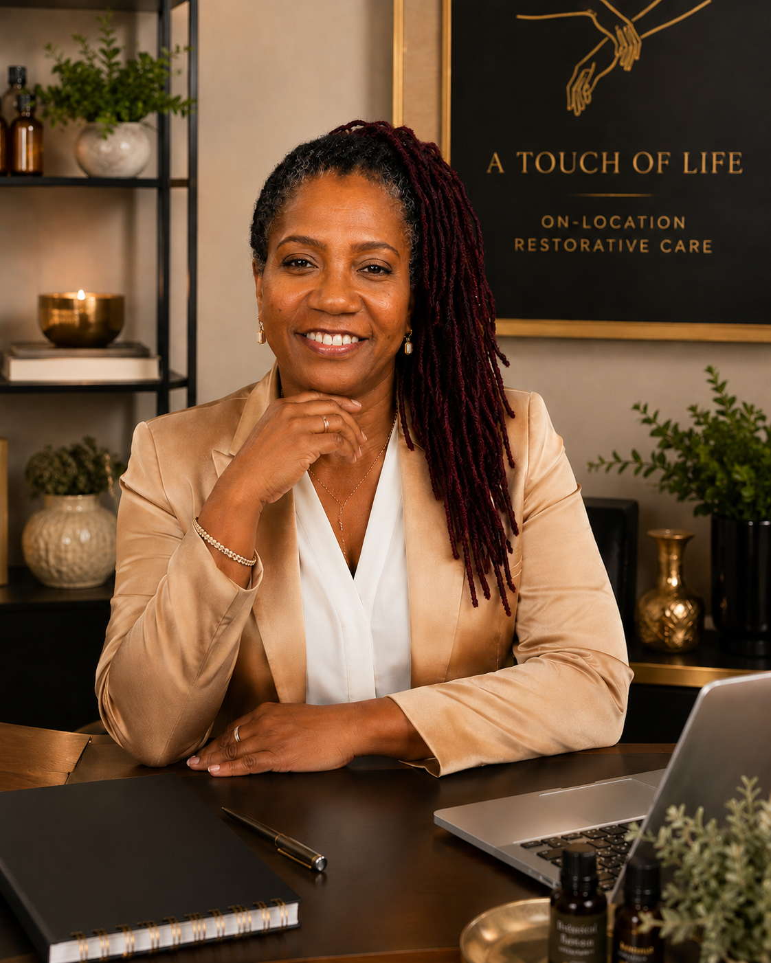 A smiling woman with dark skin and red dreadlocks wearing a beige blazer and white blouse sitting at a desk in a modern office or wellness space. Behind her are decorative plants and a black sign with gold lettering that reads 'A Touch of Life' and 'On-Location Restorative Care.'