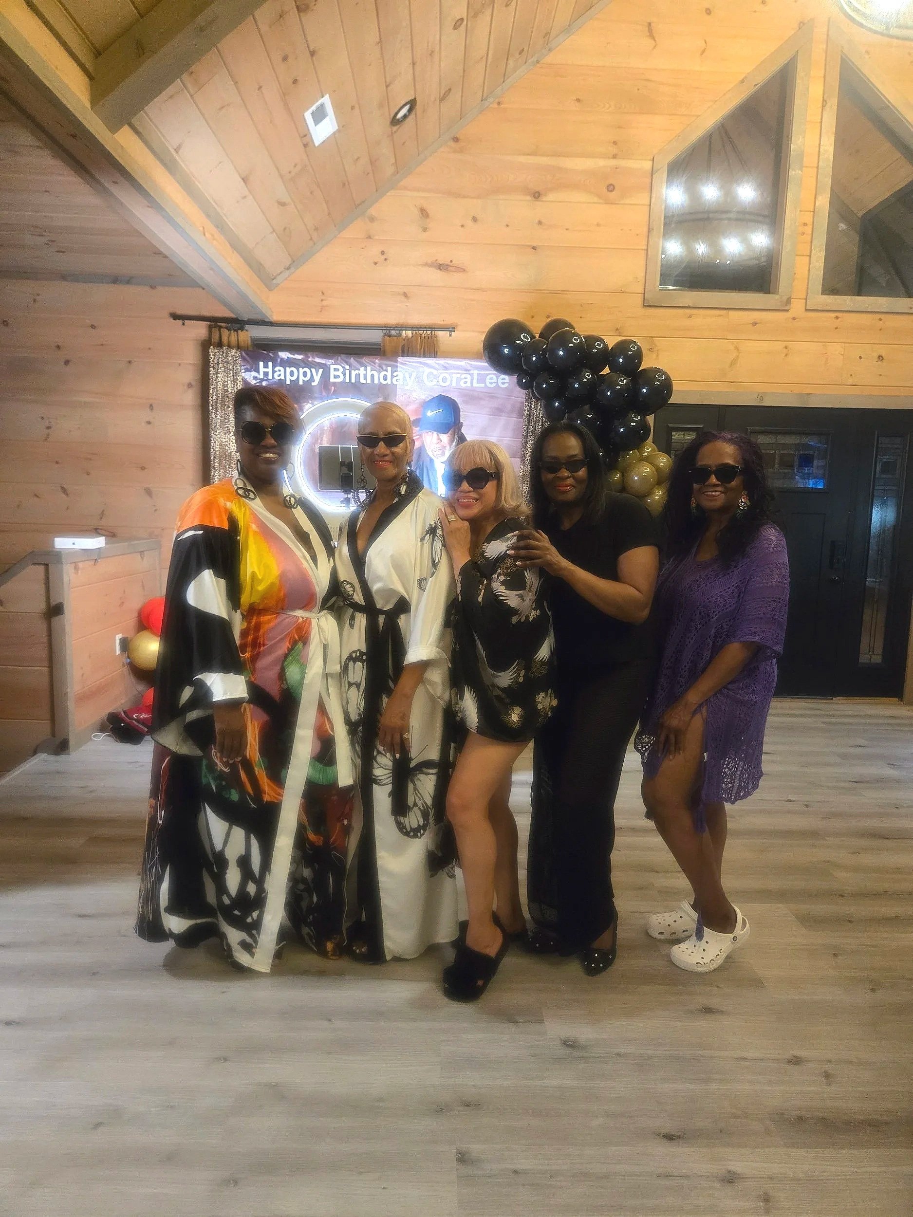 Four women standing together at a birthday celebration, smiling and wearing sunglasses. Behind them is a banner with the text 'Happy Birthday Cora Lee' and bubble balloons in black and gold.