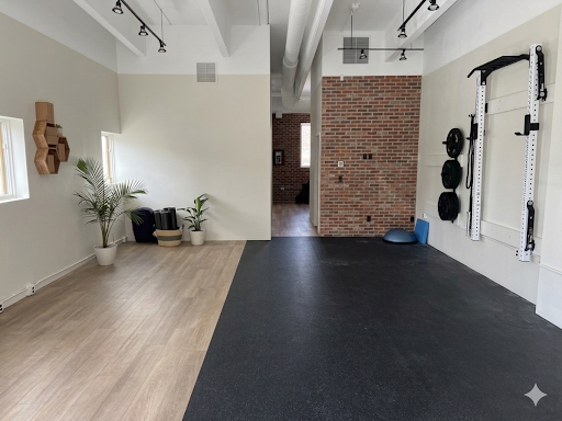 Empty workout space with black rubber flooring and white walls, featuring wall-mounted exercise equipment, potted plants, and wooden wall shelves.