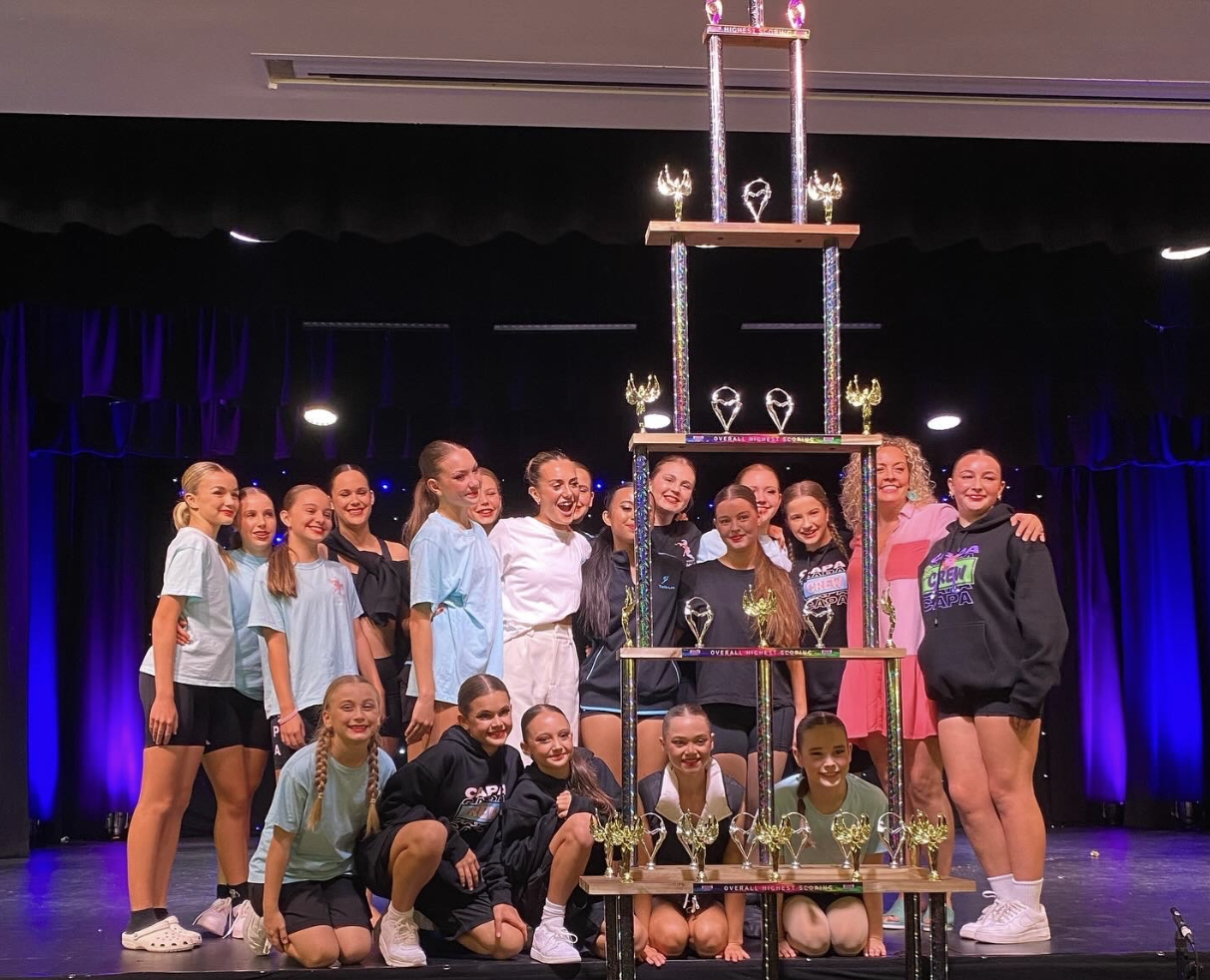 A cheerleading team of young girls and their coaches standing on a stage behind a large trophy, smiling after a competition.