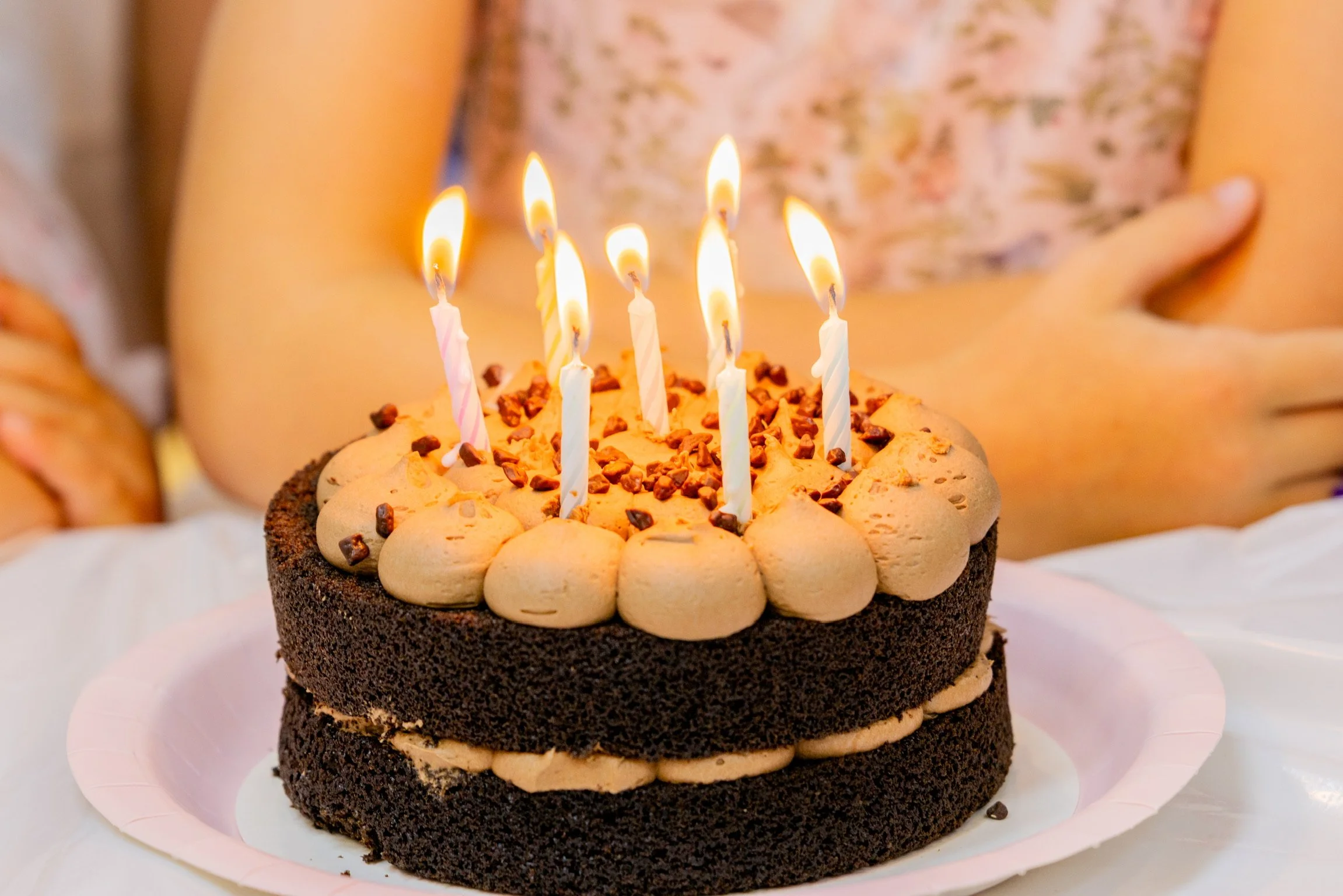 A birthday cake with six lit candles, decorated with cream and chocolate sprinkles, on a pink plate, with a person in the background.