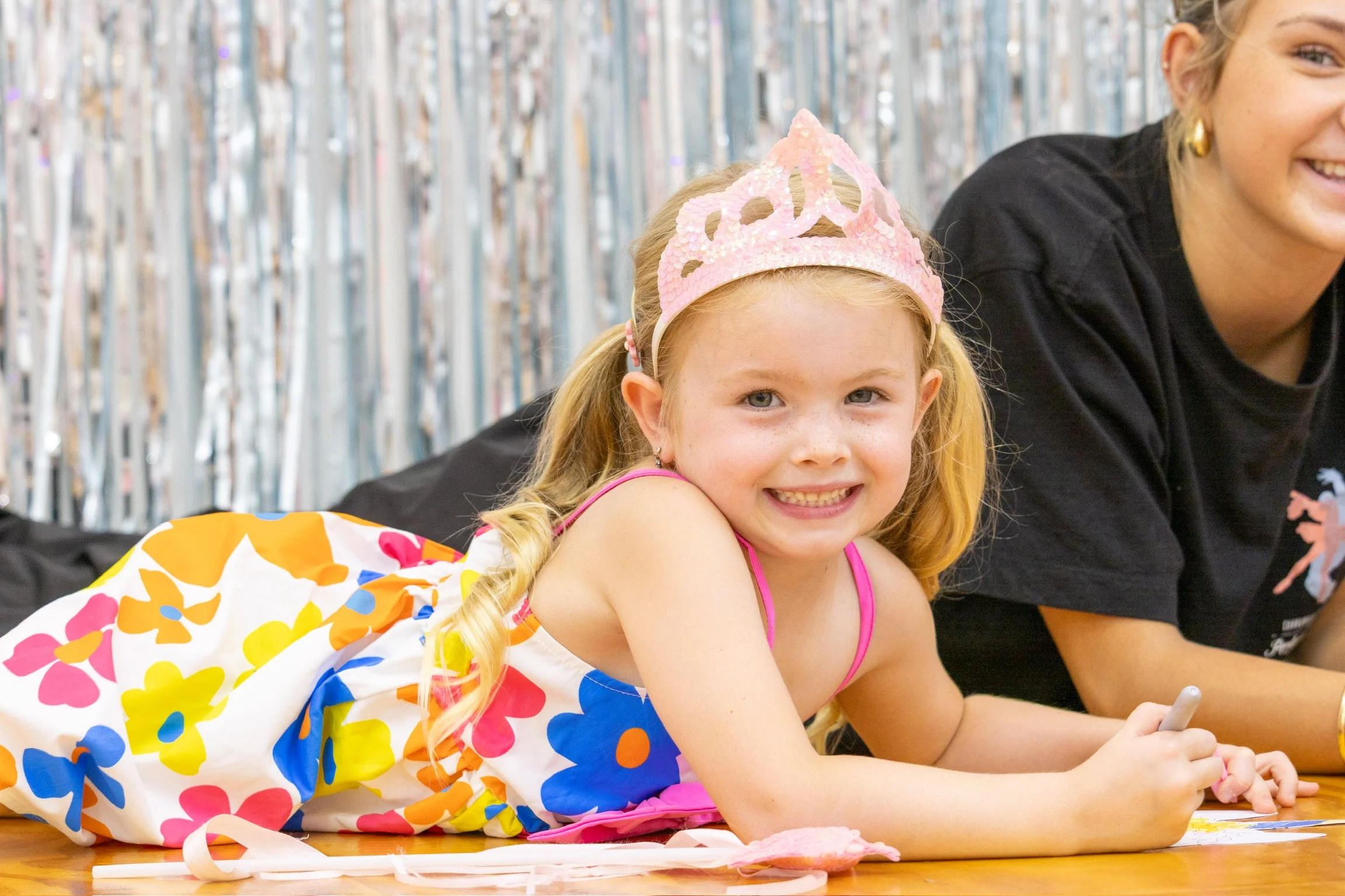 A young girl with red hair, blue eyes, and freckles lying on her stomach on a wooden floor, wearing a colorful floral dress and a pink tiara, smiling at the camera. An adult woman is partially visible next to her.