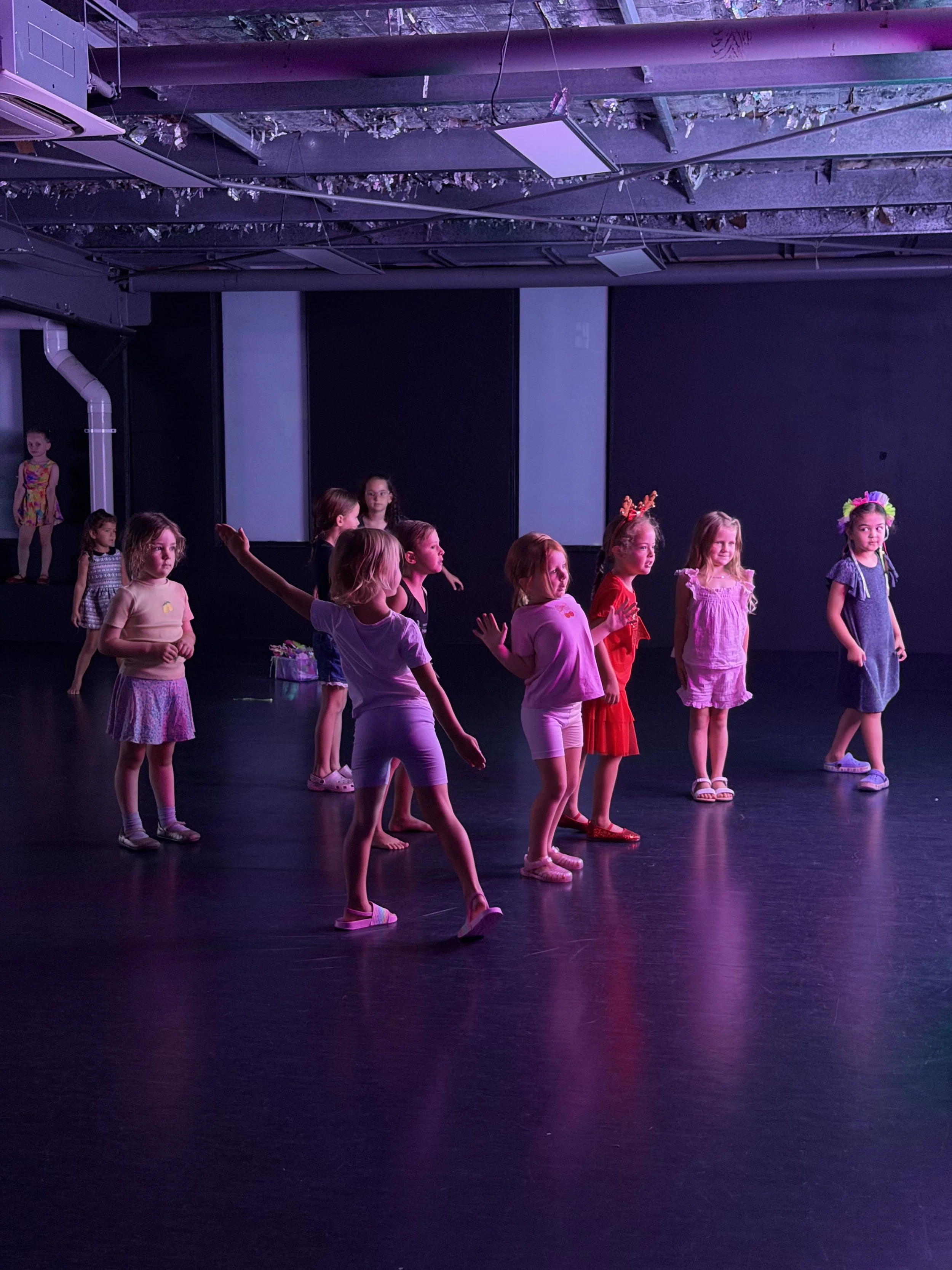Group of young girls practicing dance or performing on stage in a dimly lit space with purple lighting.