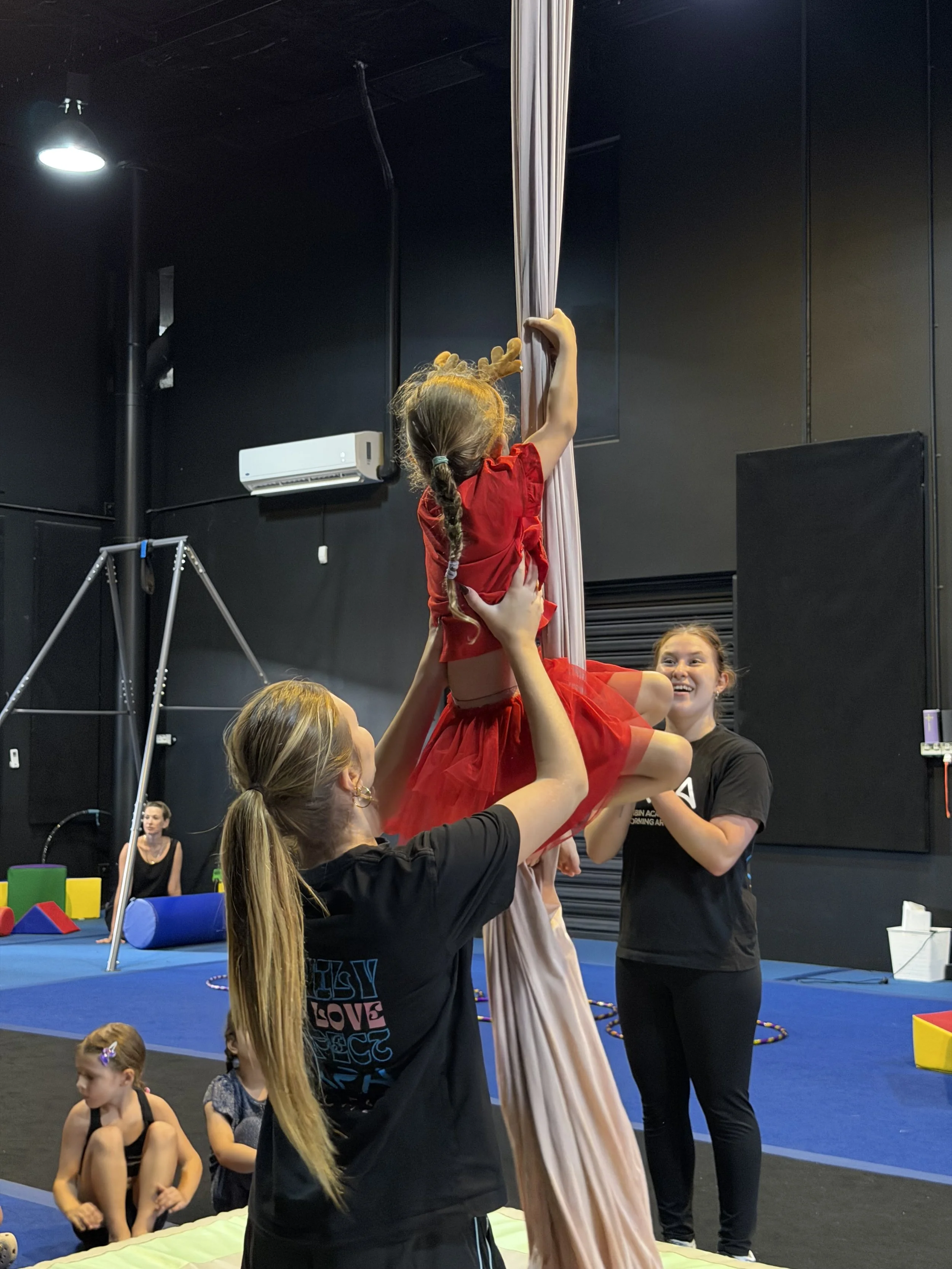 A girl performing aerial silk acrobatics, supported by two women during a class or practice session at a gym with black walls, other children and equipment visible in the background.