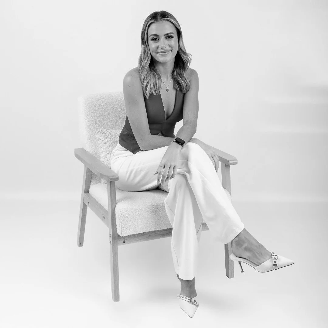 A woman sitting on a wooden chair with a cushioned seat against a white background. She has shoulder-length hair, is smiling, and is dressed in a sleeveless top, white pants, and high heels. She is wearing a fitness tracker on her left wrist and a necklace.
