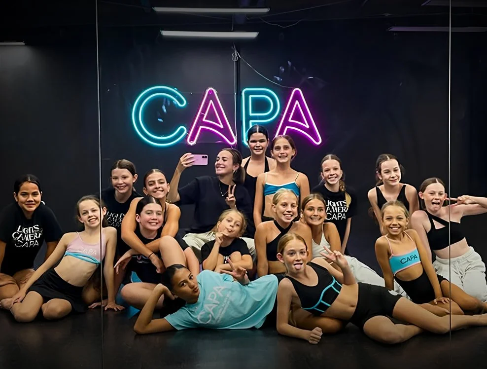 Group of young dancers posing in front of a mirror with a neon sign reading 'CAPA' in pink and blue lights at a dance studio.