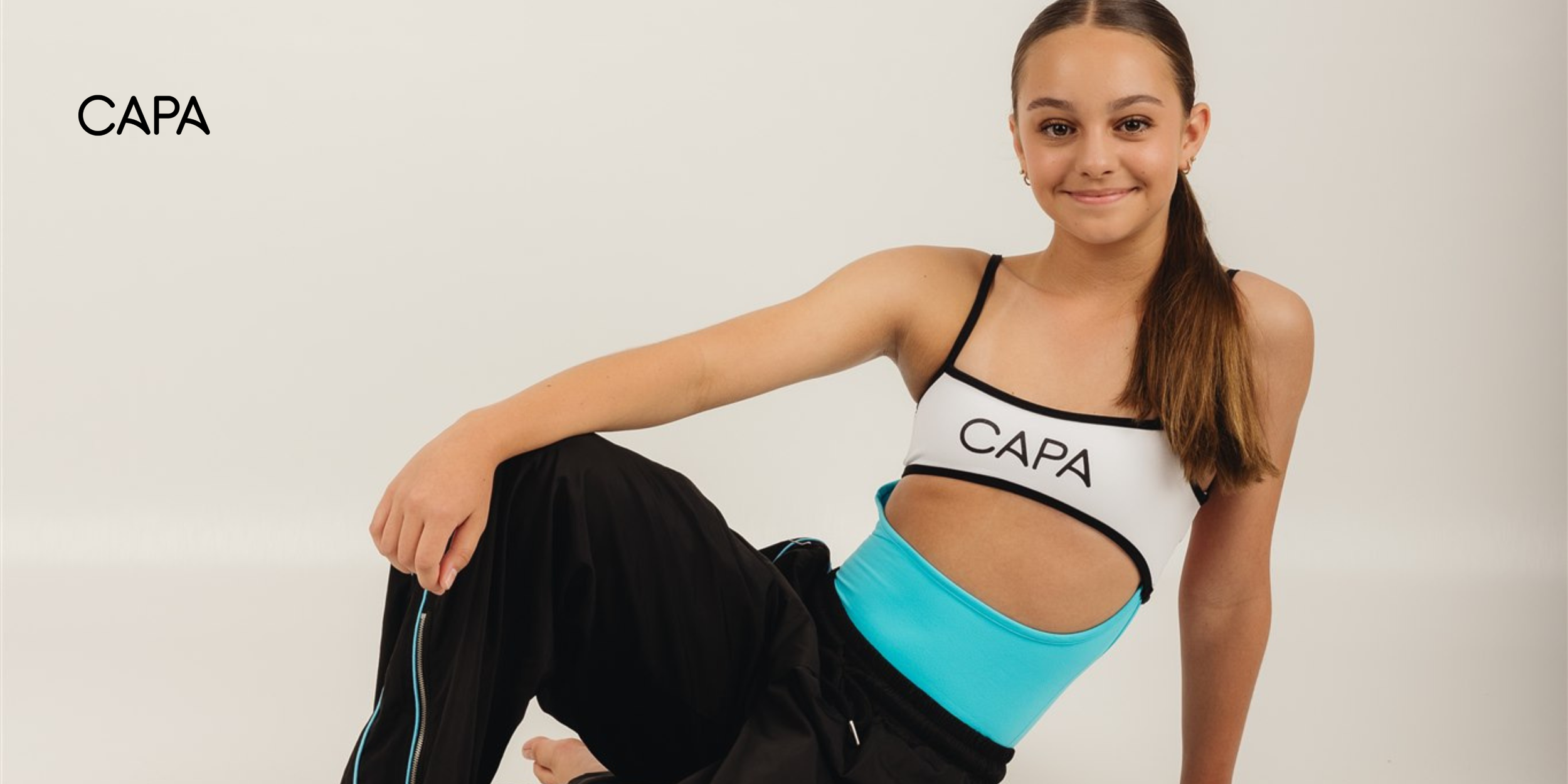 A young woman wearing a sports bra with the text 'CAPA' and black straps, along with black pants with blue accents, sitting on the floor against a plain white background.