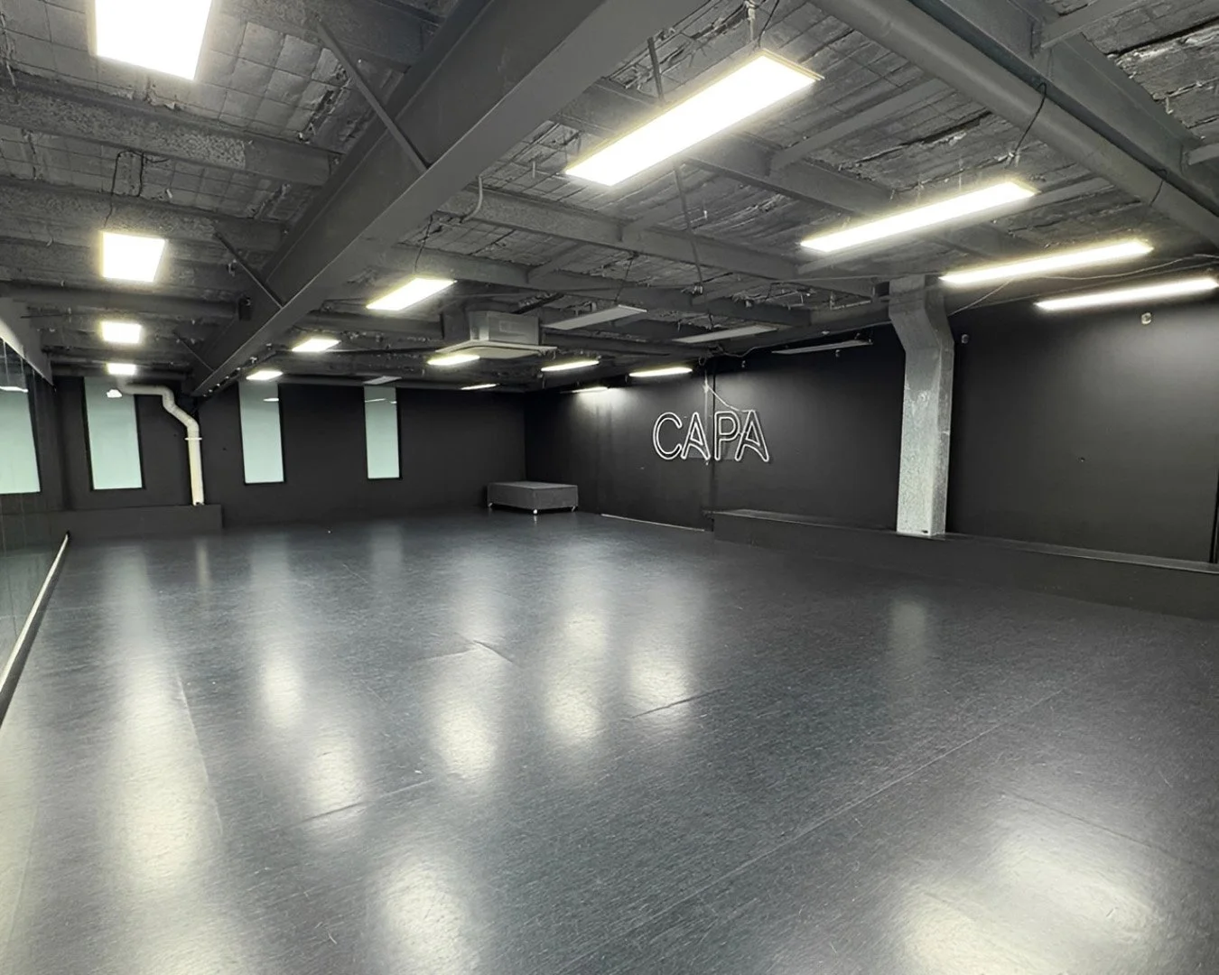 Empty dance studio with black walls, black flooring, exposed ceiling piping, and fluorescent lighting. The word 'CAPA' is displayed on the wall in illuminated letters.