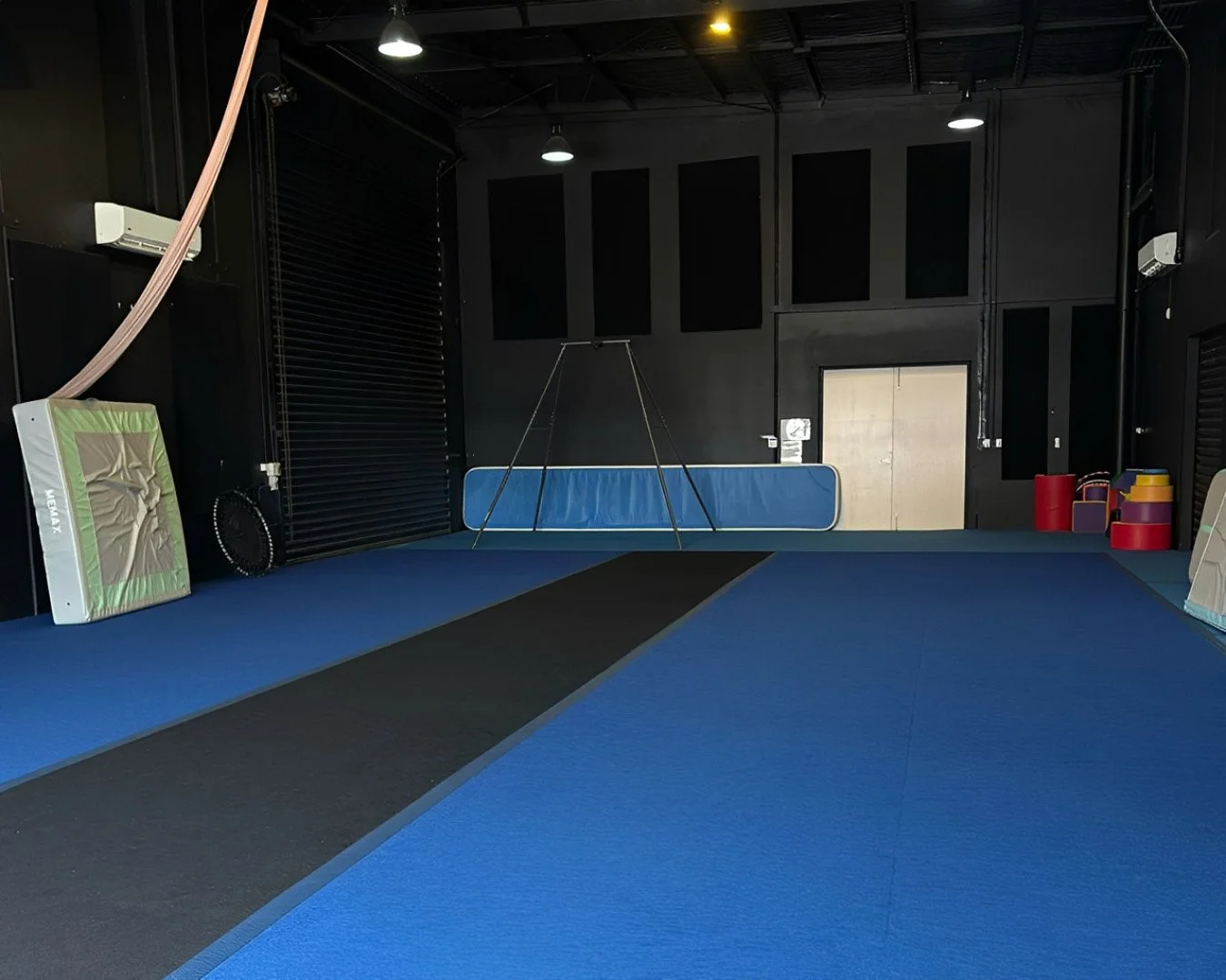 Interior view of a martial arts or gym training room with blue and black mats, padded equipment, and various training gear against black walls, including a large black fan and colorful foam rollers.