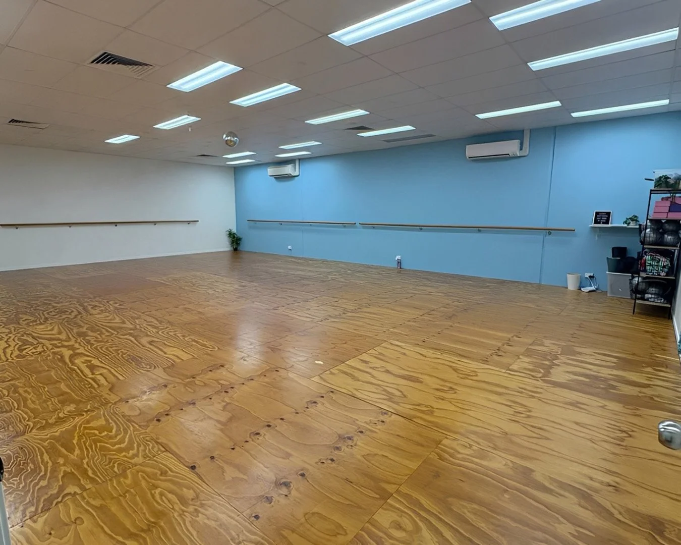 Empty dance studio with wooden floor, blue and white walls, horizontal ballet bars, and ceiling lights.