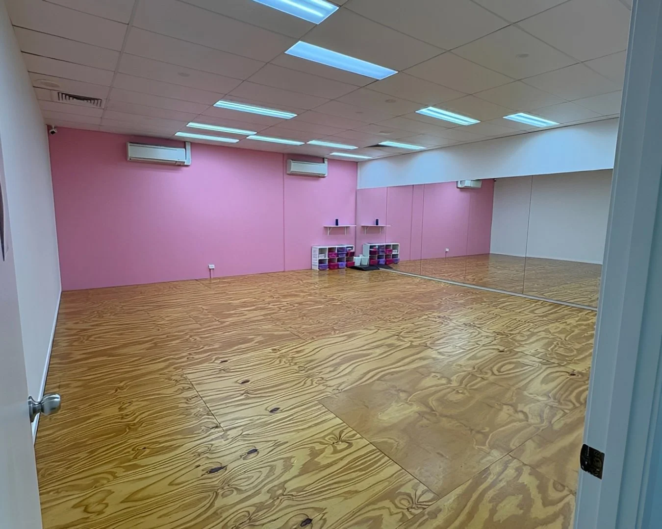Empty dance studio with wooden floors, pink walls, a mirror on one wall, and a small yoga mat and shelves with exercise equipment.