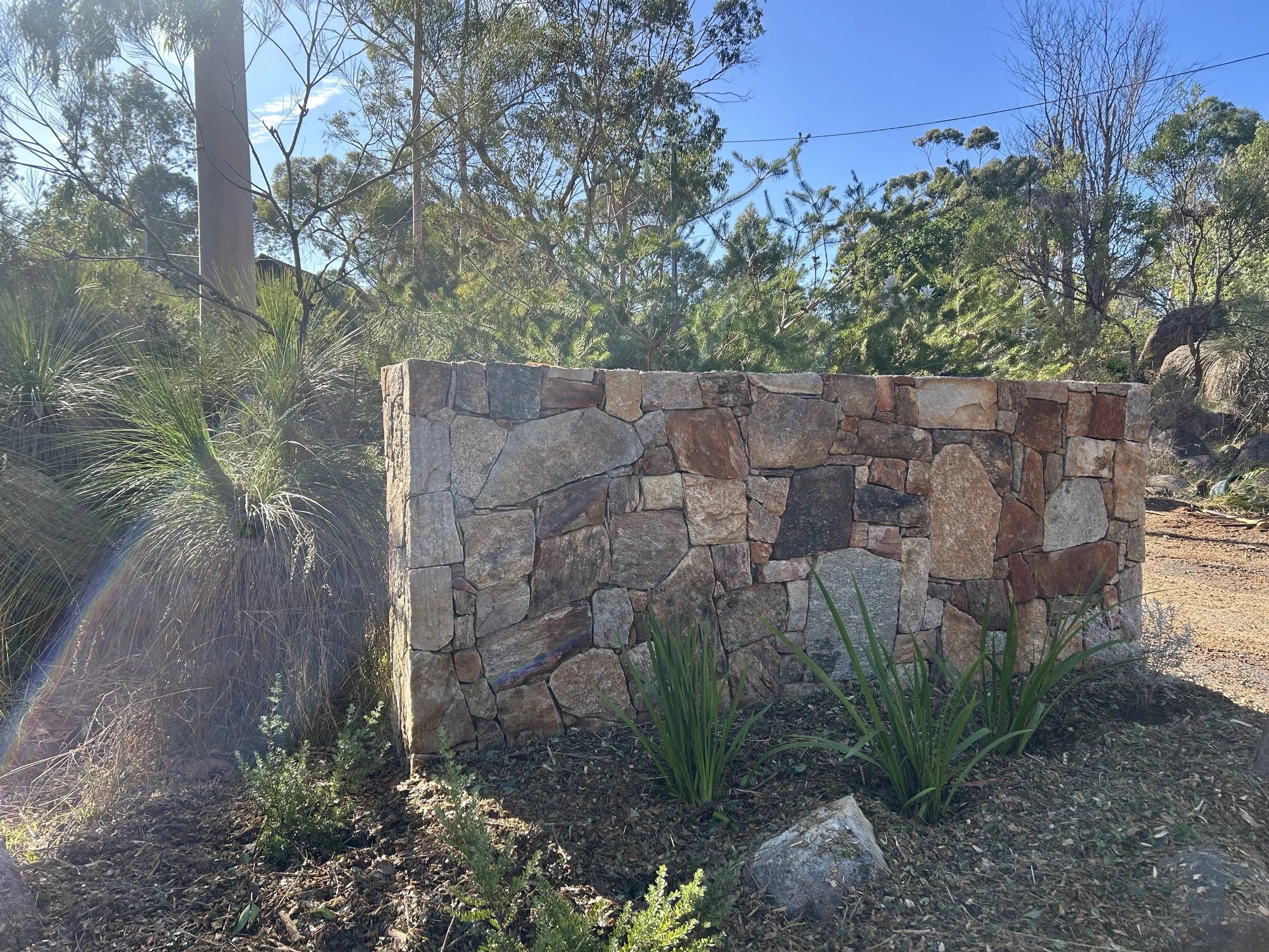 A landscape with a stone wall, various green plants and trees, and a background of a clear blue sky.